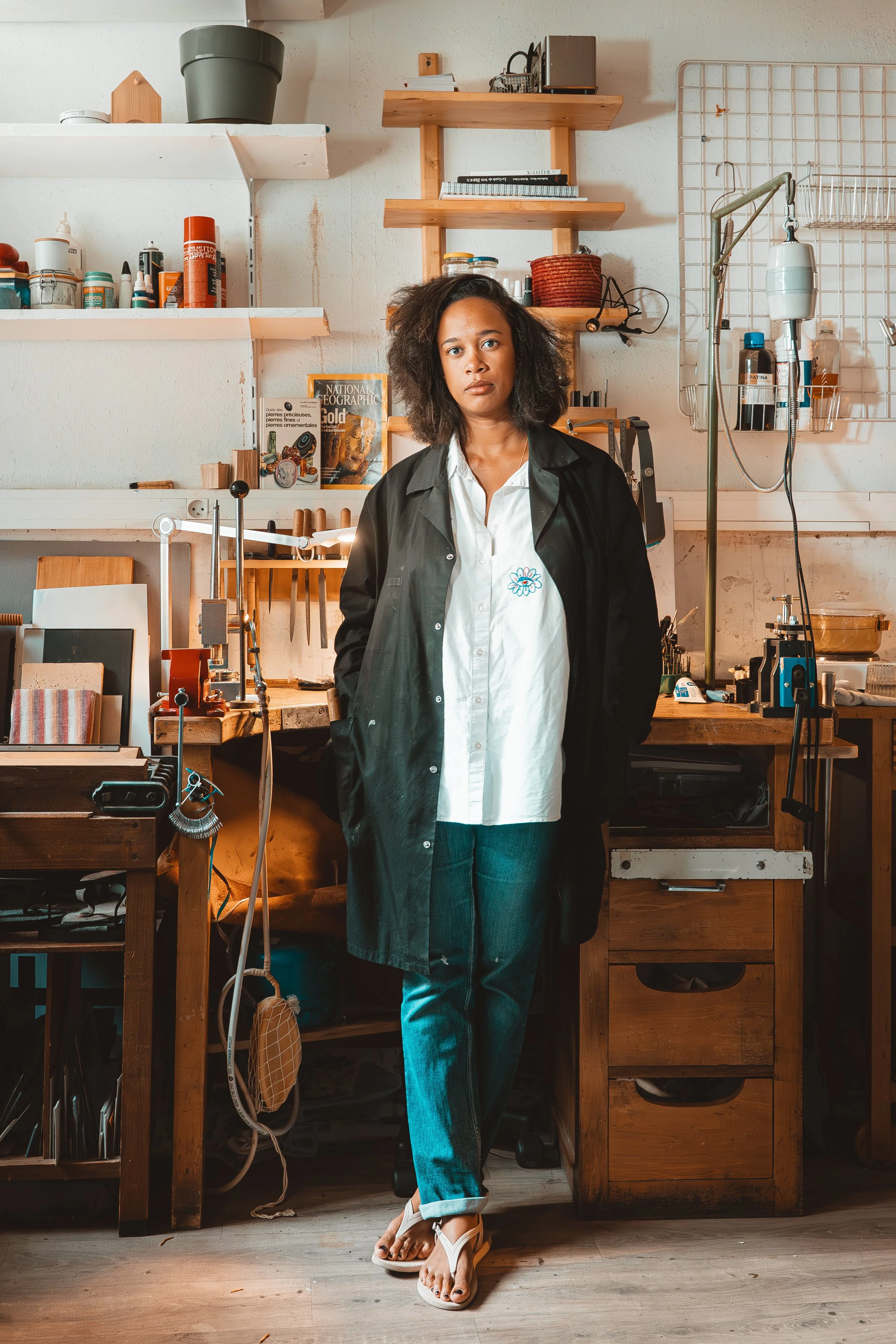 A woman standing in a workshop or art studio, surrounded by shelves and various supplies, wearing a black coat over a white shirt and jeans.