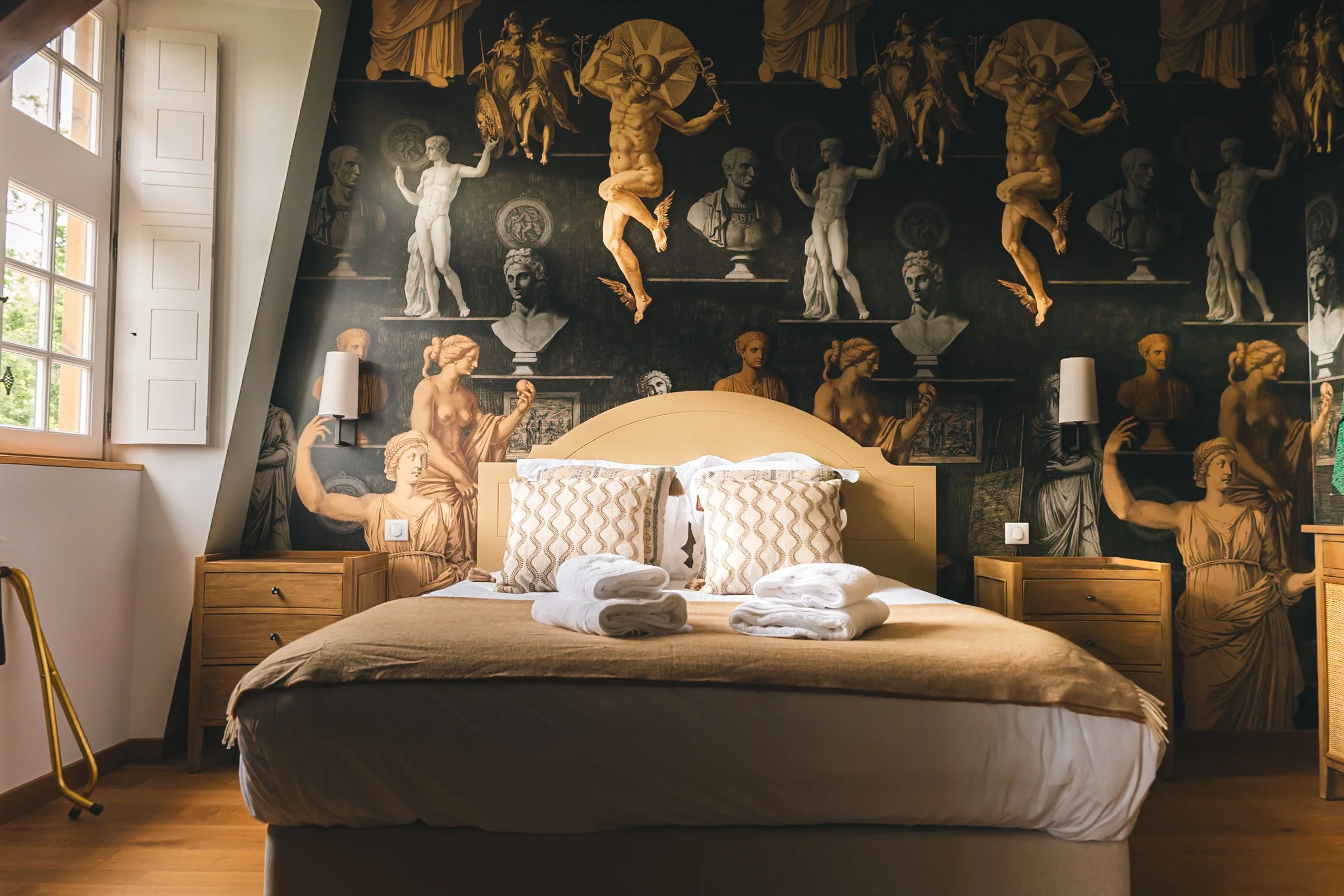 A bedroom with a large mural featuring classical sculptures and figures on the wall behind the bed. The bed has pillows and neatly folded white towels on top, with bedside tables on each side. Natural light comes through a window on the left.