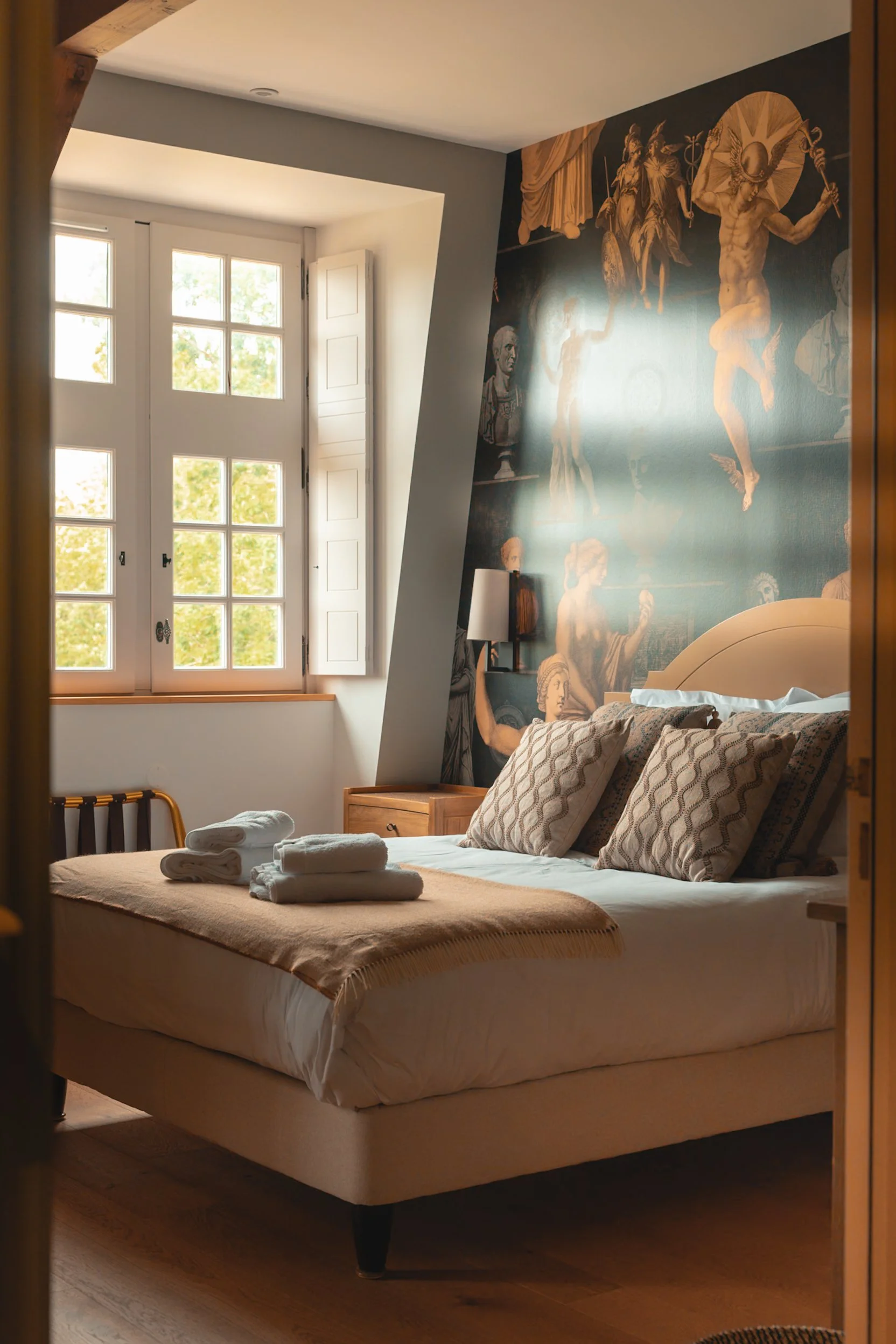 A cozy bedroom with a large window, a bed with patterned pillows and folded towels on top, a wooden nightstand with a lamp, and a wall mural depicting mythological figures.