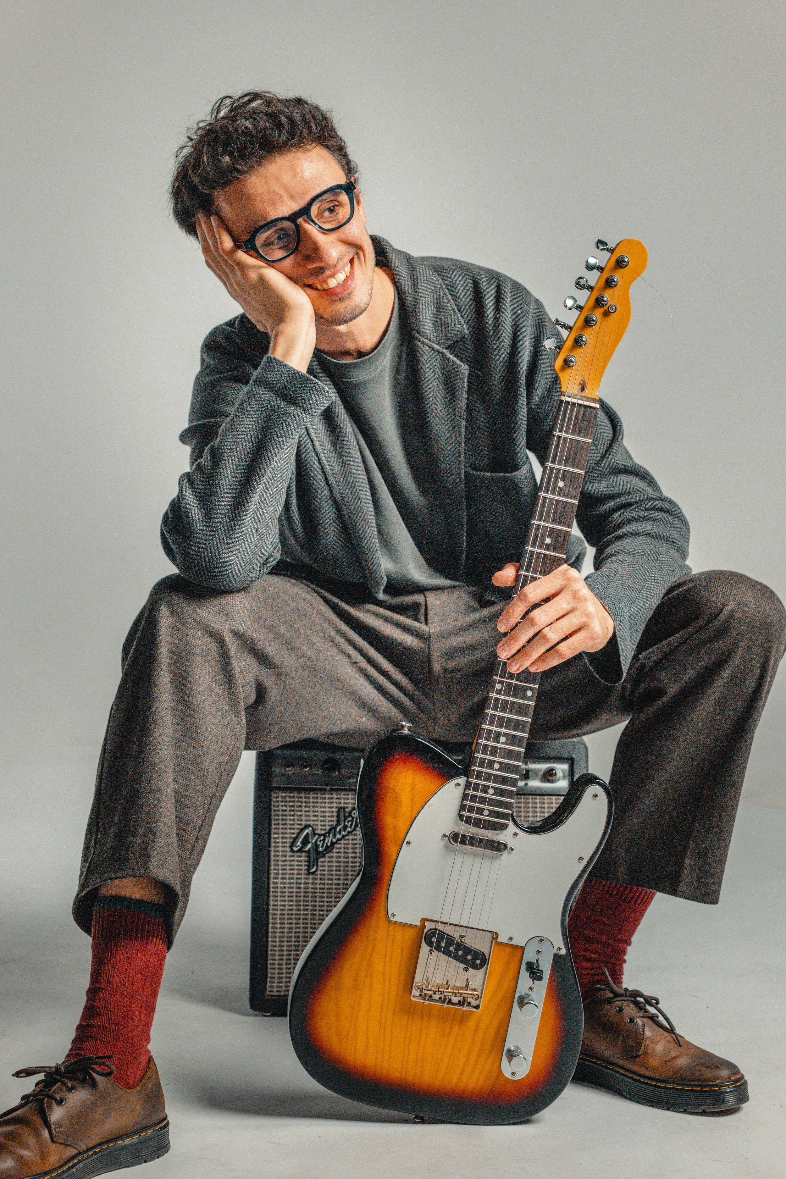 A man with glasses and curly hair sitting on a guitar amplifier, holding an electric guitar, smiling and resting his head on his hand, dressed in casual vintage clothing.