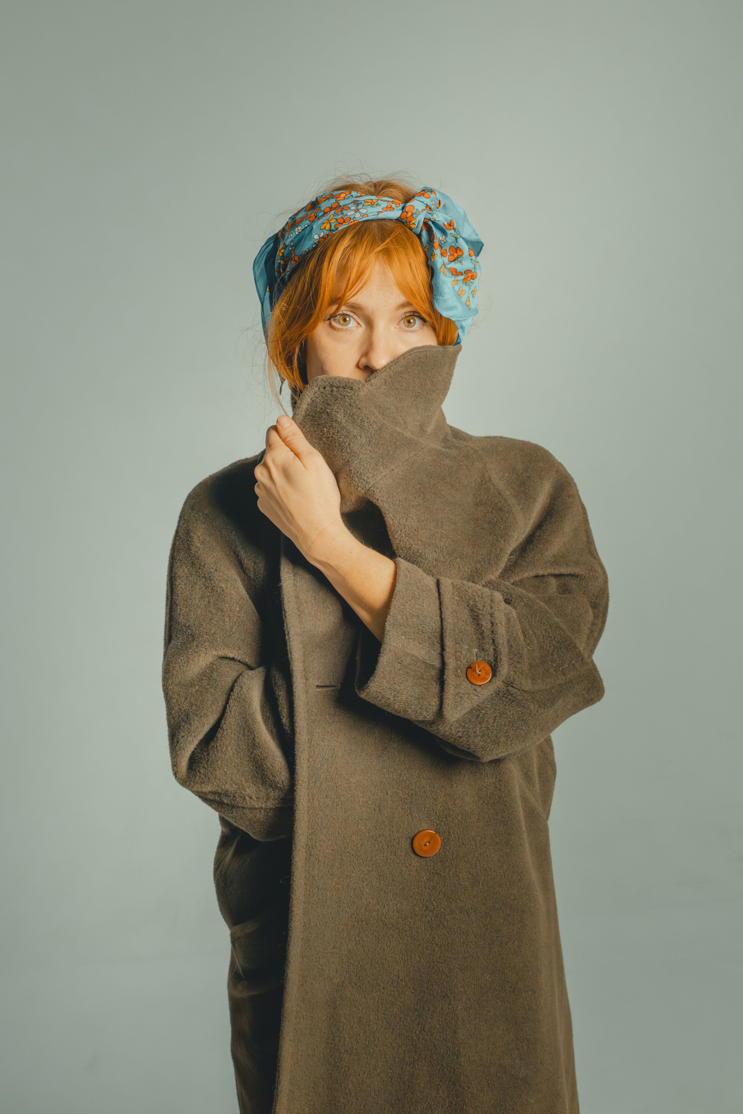A woman with red hair wearing a blue headscarf with orange floral patterns, holding a brown coat collar up to her face, with a neutral background.