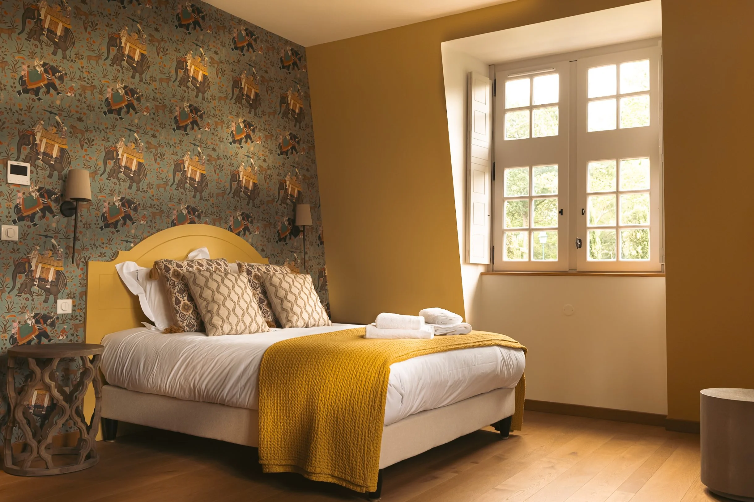 A cozy bedroom with a bed covered in white sheets, a yellow blanket, and decorative pillows. The accent wall behind the bed has patterned wallpaper with elephants and foliage. There are two wall-mounted lamps on either side of the bed, and a window w