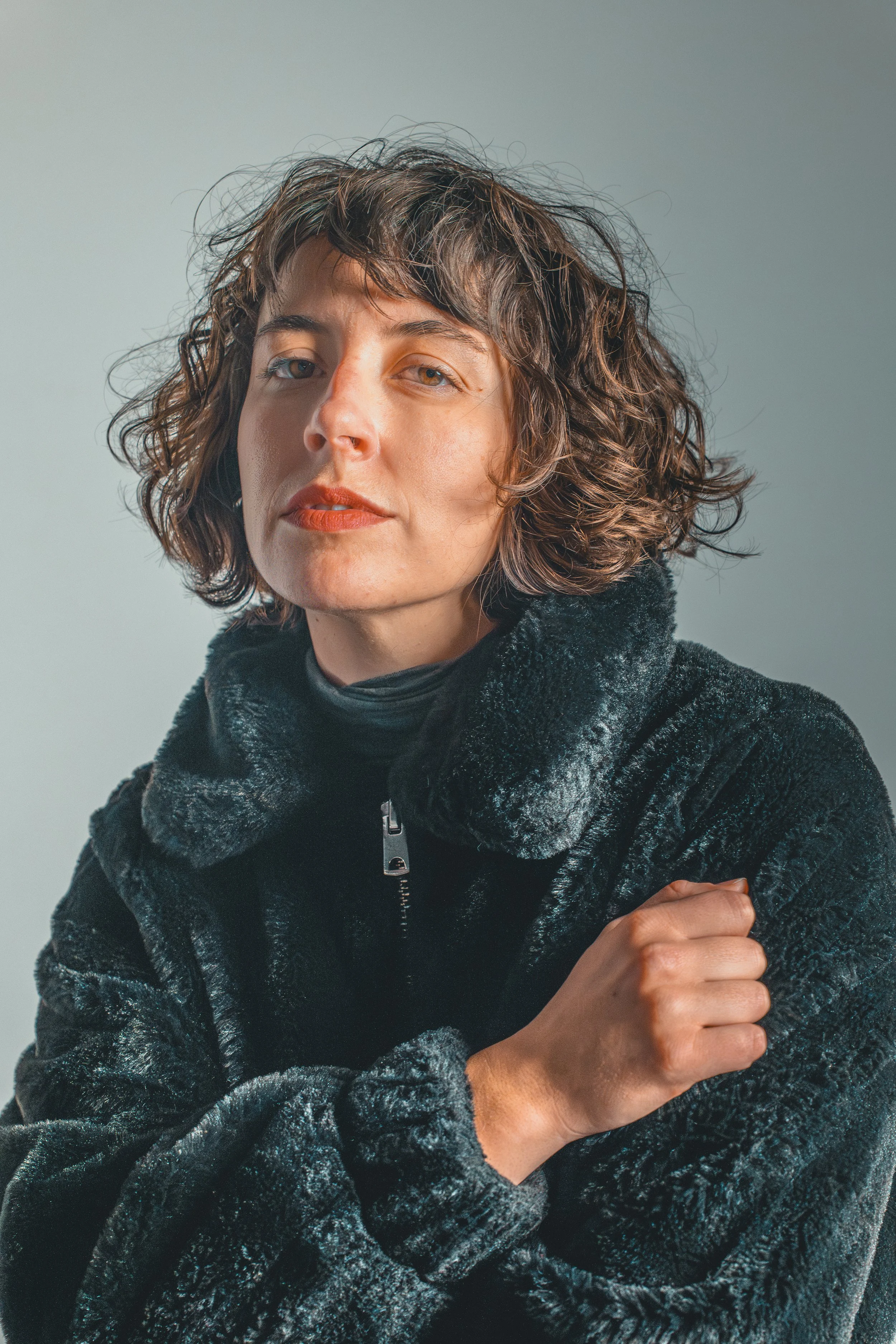 A woman with curly brown hair wearing a black, fuzzy zip-up jacket and a black turtleneck, looking confidently at the camera.