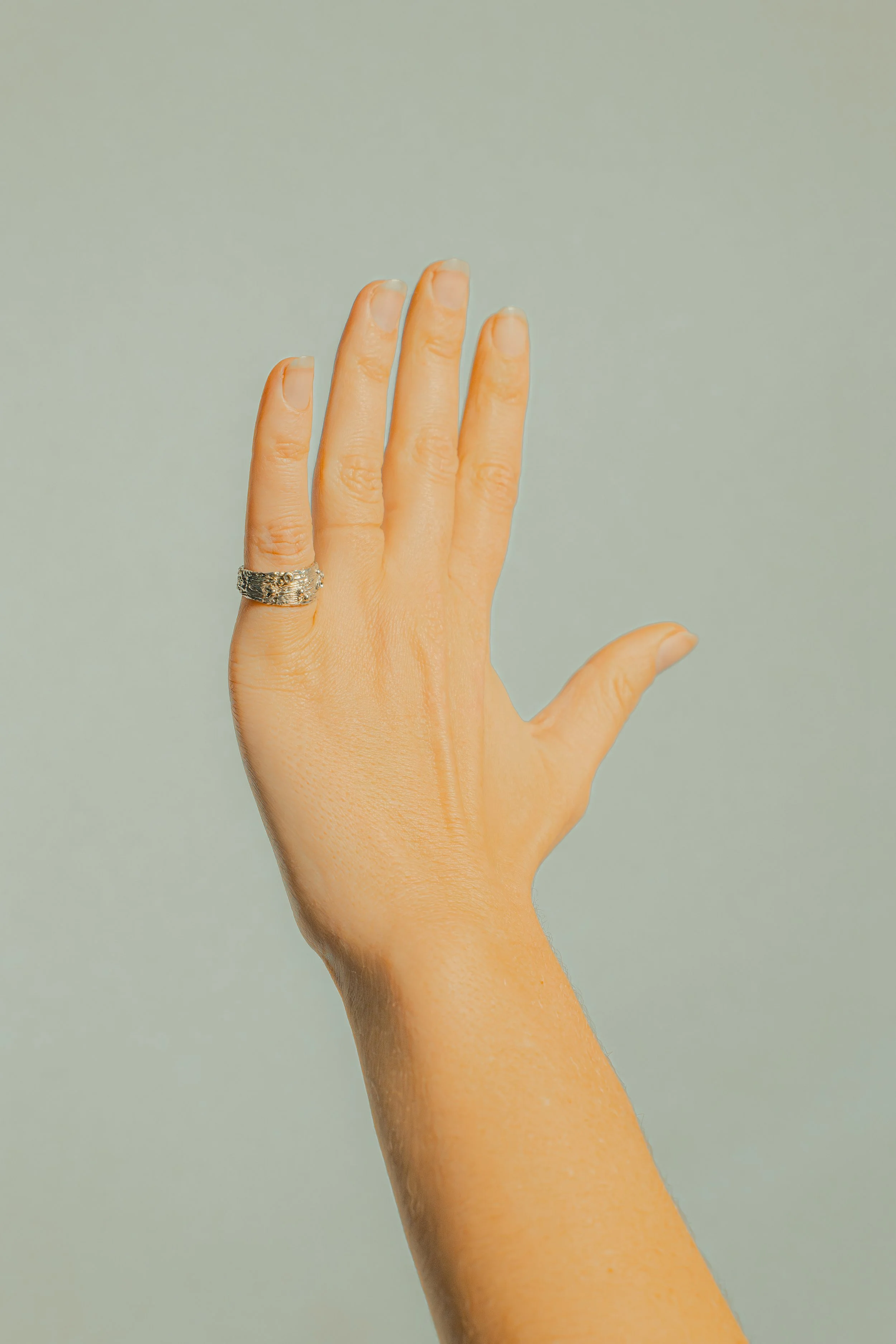 Close-up of a person's left hand with a silver ring on the middle finger, raised against a plain light-colored background.