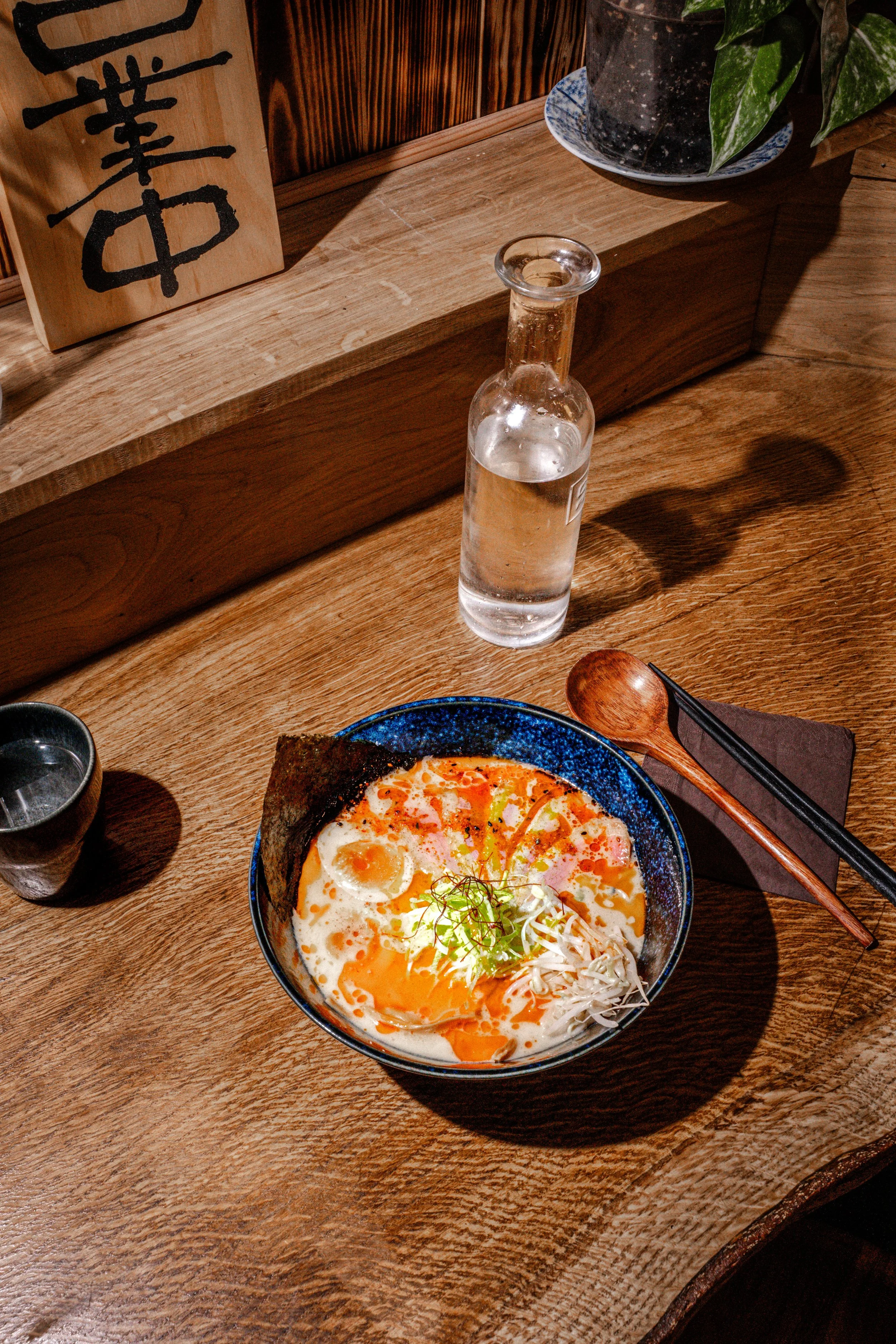 A bowl of ramen with eggs, shredded vegetables, and seaweed on a wooden table, with a small cup, chopsticks, a wooden spoon, and a bottle of clear beverage, in a cozy restaurant setting.