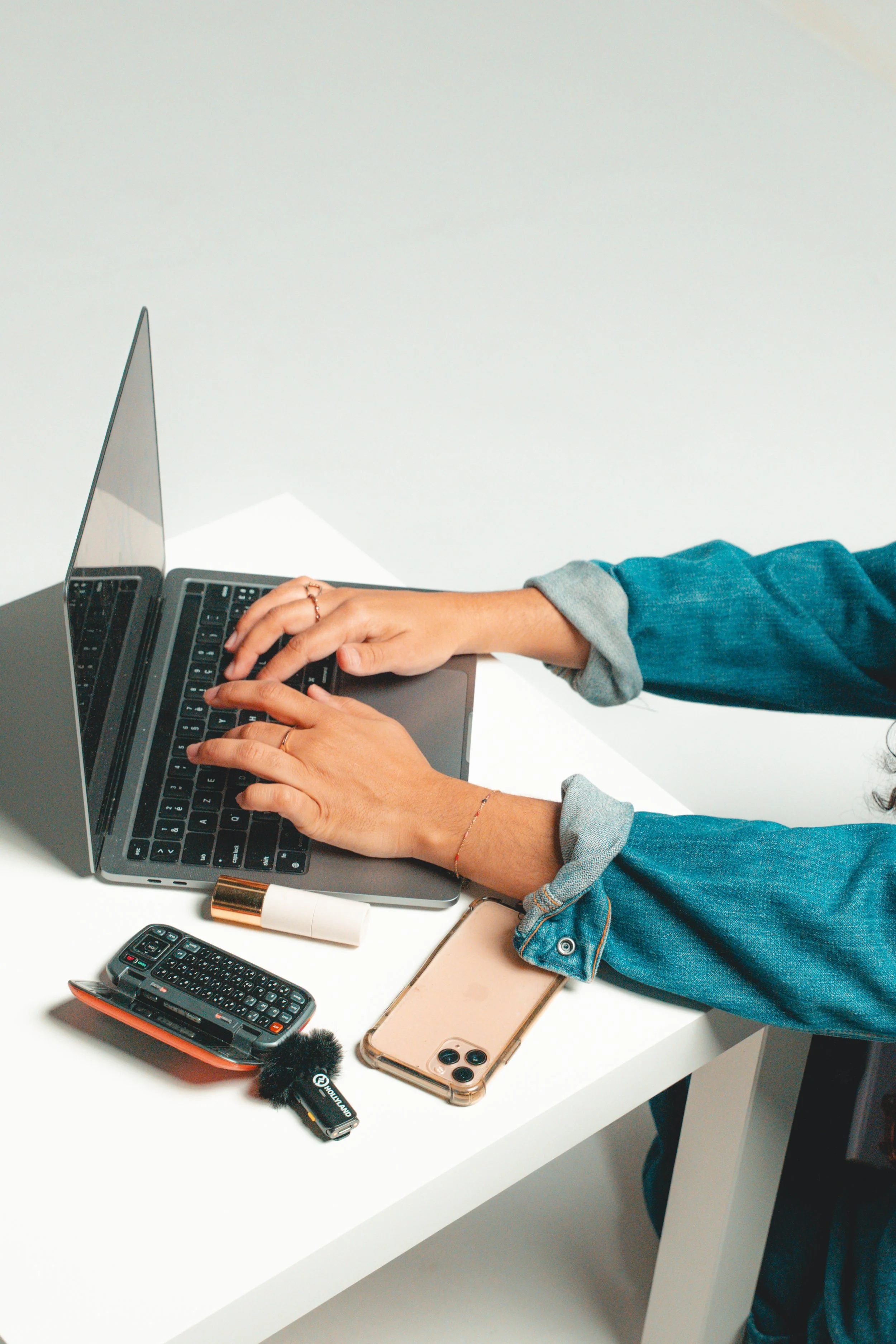 Person wearing a blue denim shirt typing on a laptop at a white desk with a smartphone, TV remote, lip balm, and small microphone nearby.