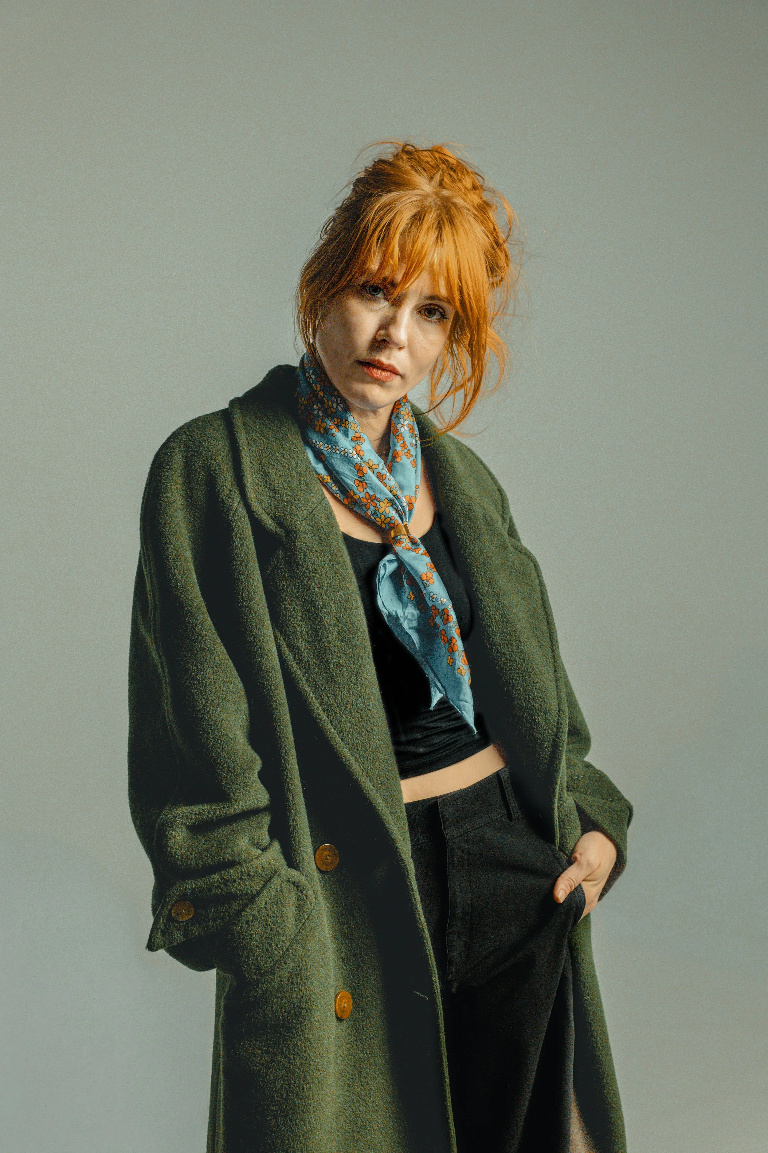 A woman with red hair wearing a green coat, a black top, and a blue patterned scarf.