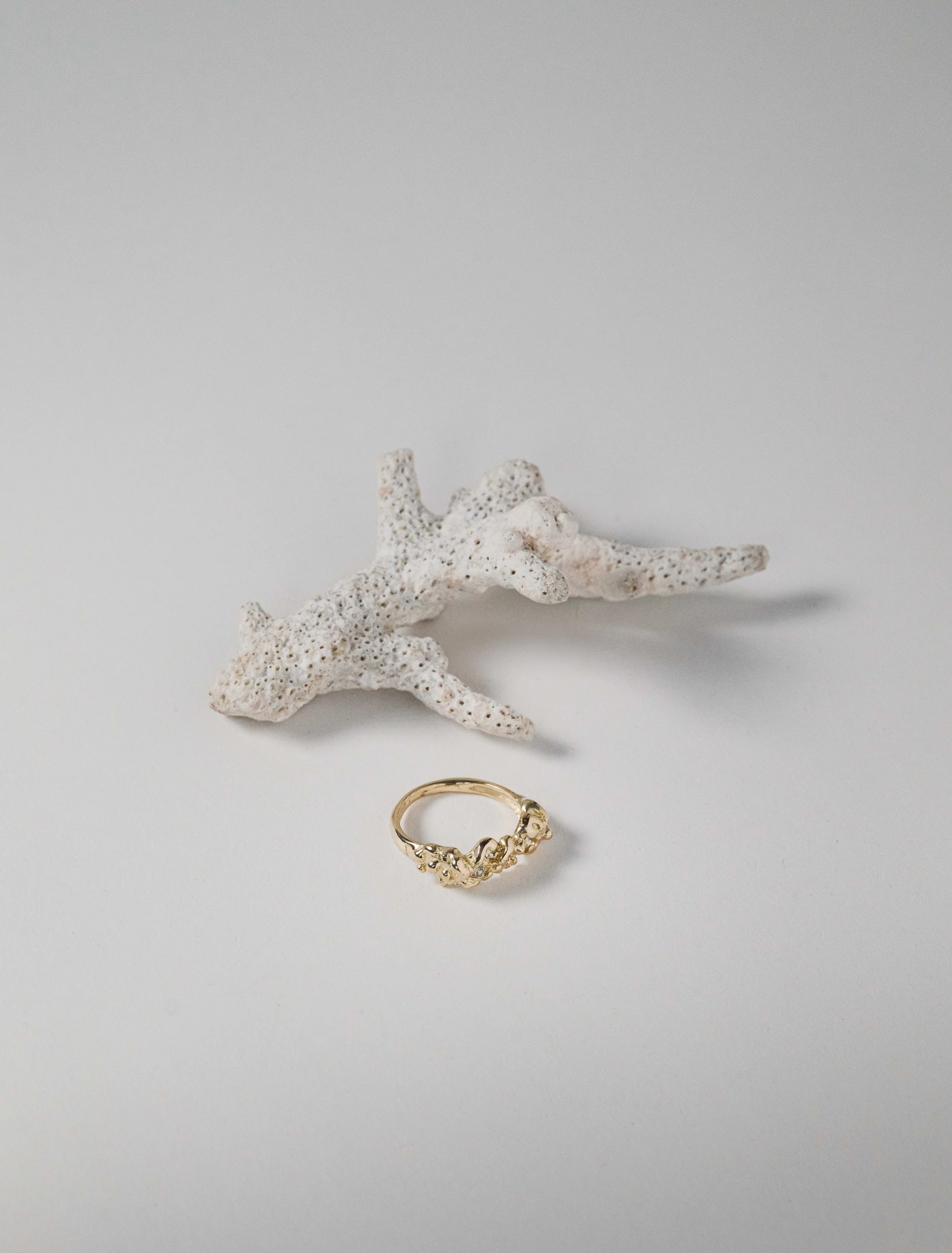 A gold ring with small decorative elements, placed on a white surface with a piece of white coral behind it.