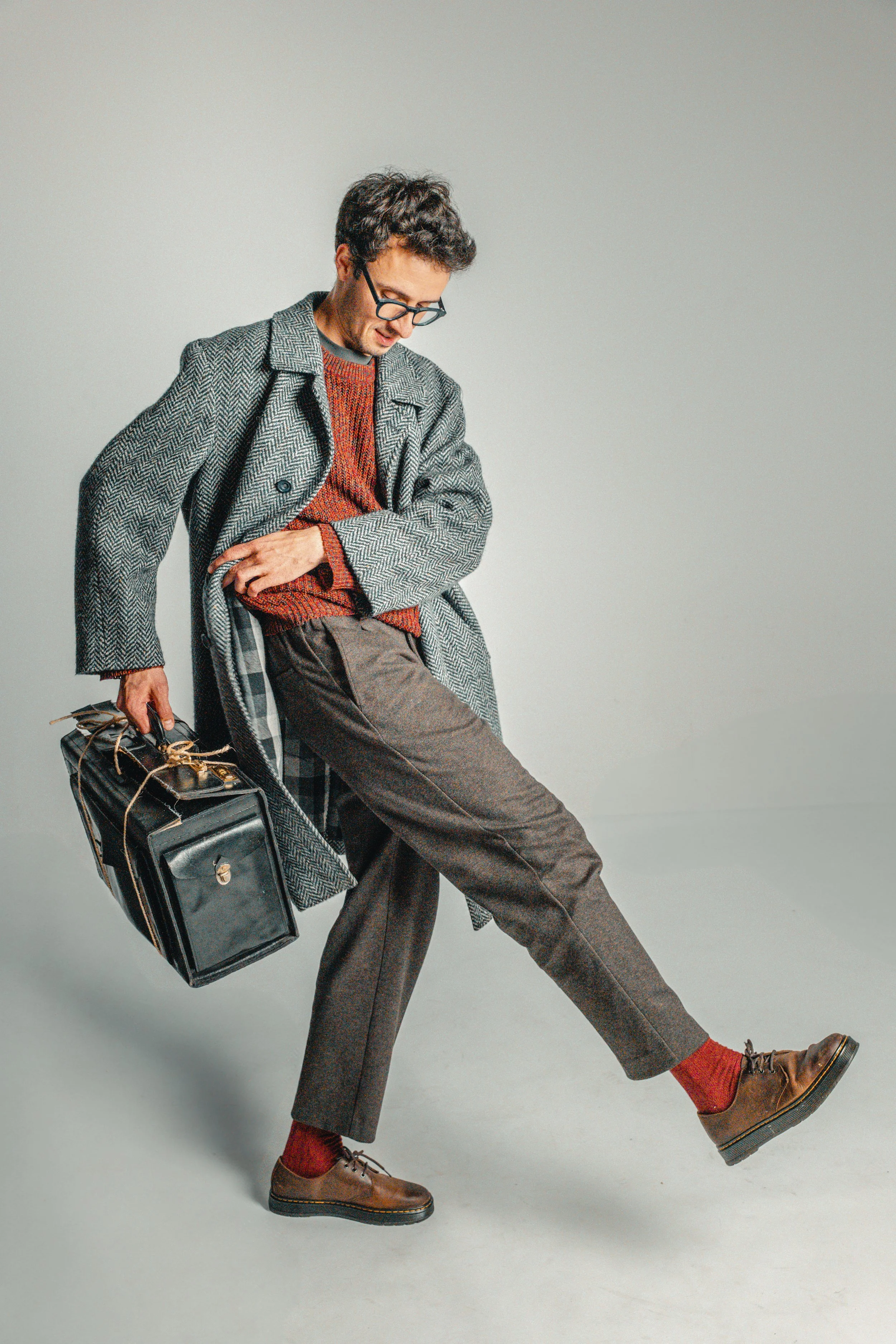 Man with glasses and dark curly hair in vintage clothing holding a briefcase, lifting his left leg in a playful pose against a plain gray background.