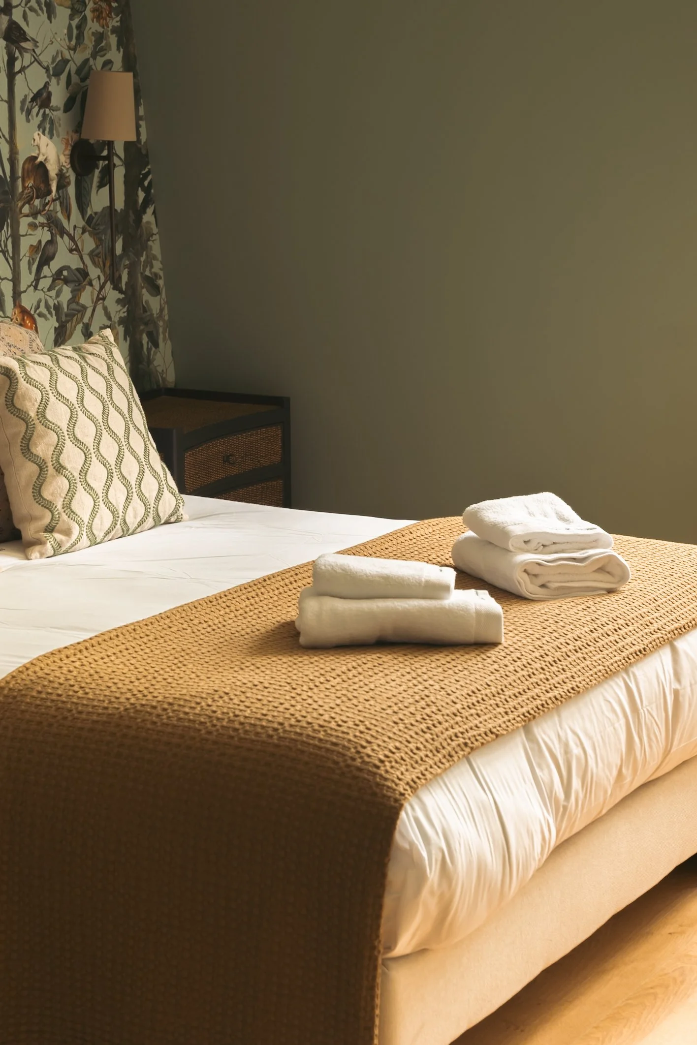 A neatly made bed with white linens, a beige bed skirt, and a textured tan bed runner. Towels and washcloths are folded on the bed. The headboard features a decorative pillow with a leaf pattern, and the background includes a floral wallpapered wall 