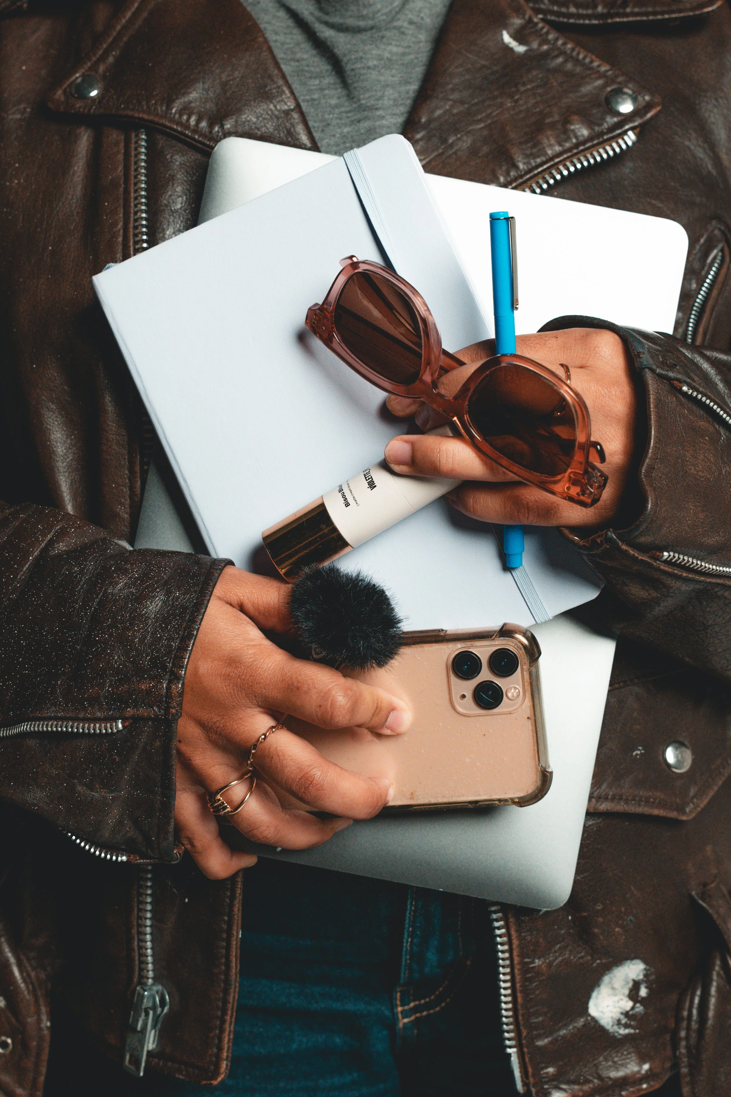 Close-up of a person holding a smartphone, sunglasses, and a tube of cream, with a laptop and notebooks inside a leather jacket.