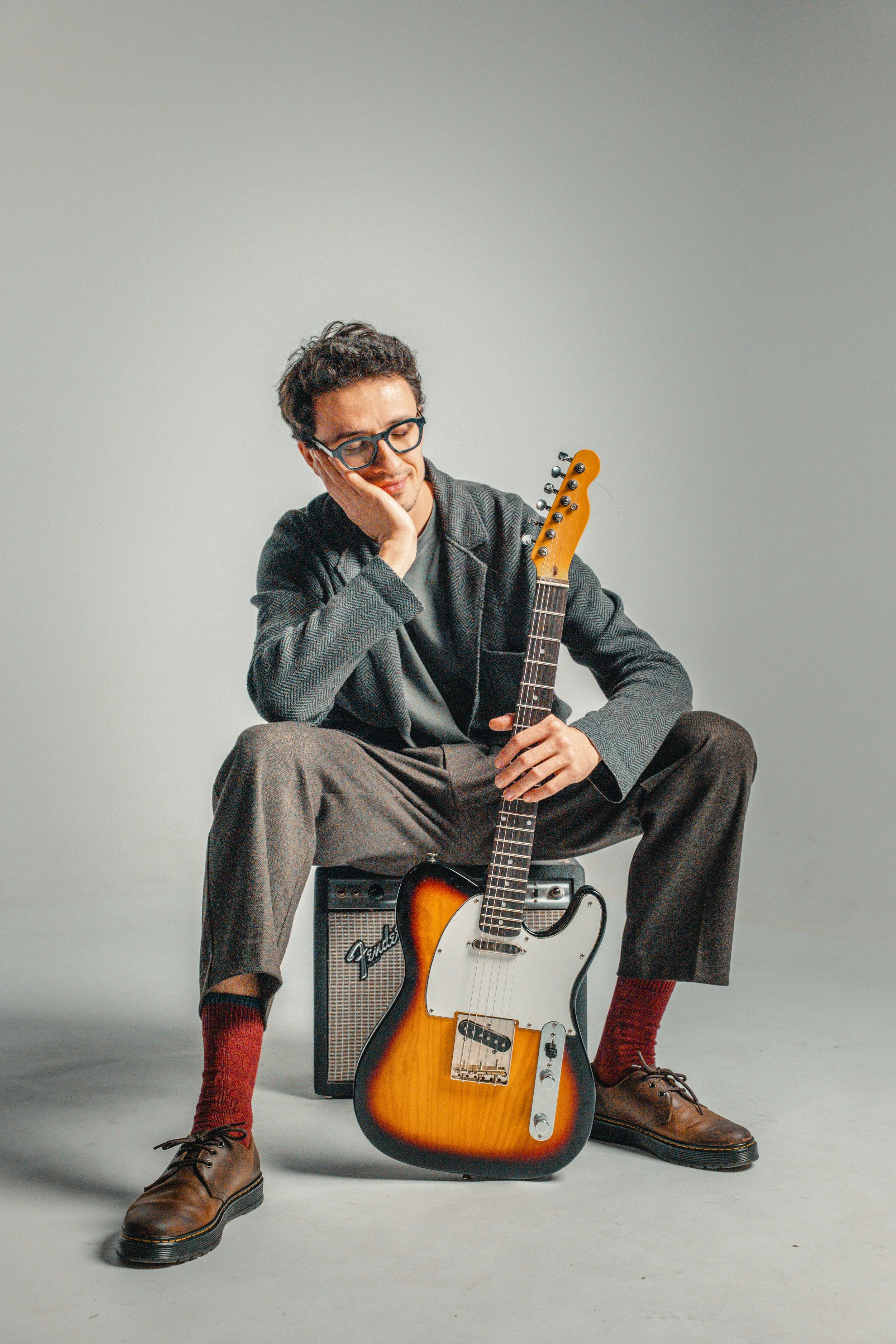 Young man sitting with headphones on, resting his head on his hand, holding an electric guitar, with a small amplifier nearby, in a studio with a plain background.