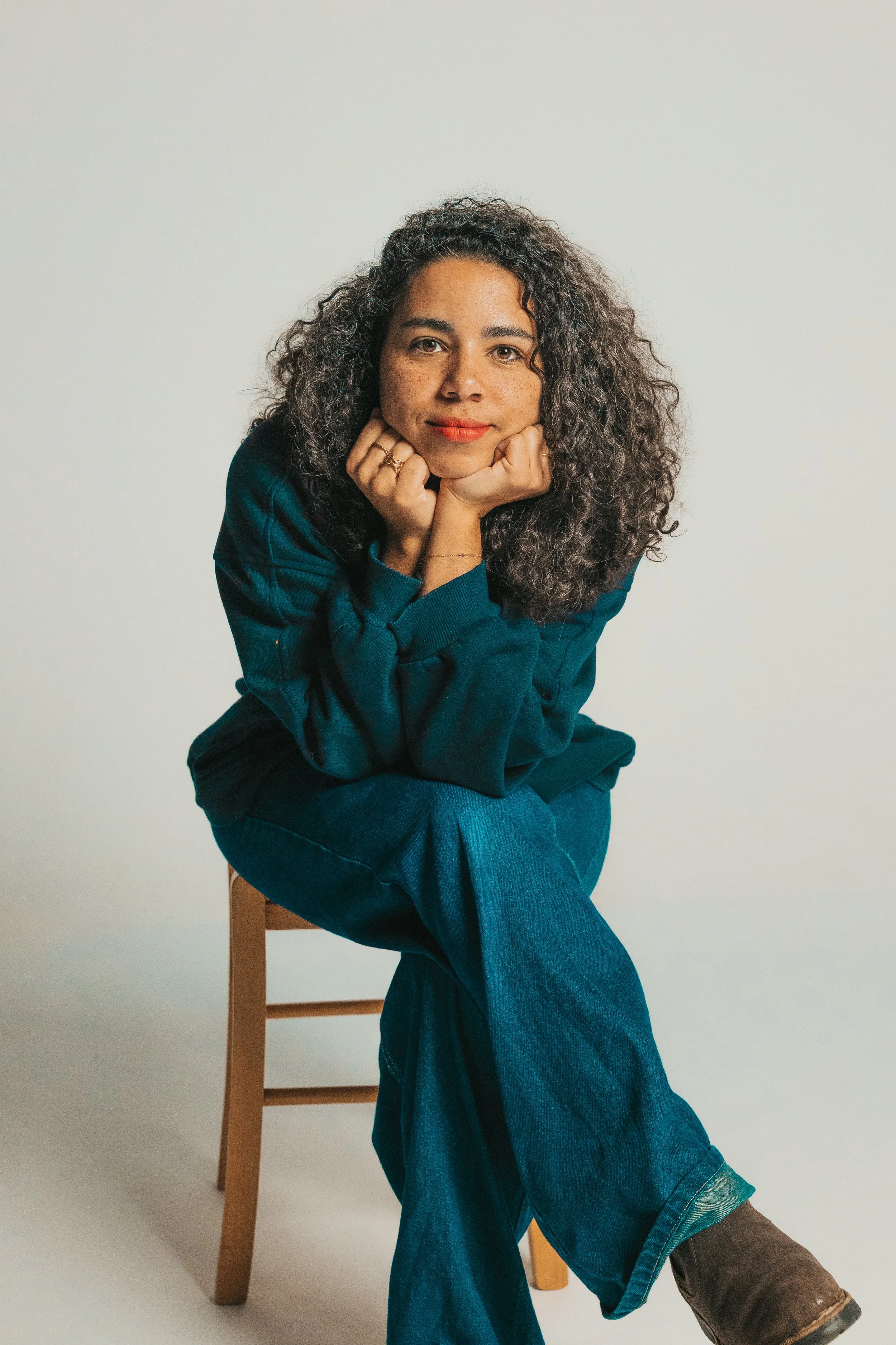 A woman with curly hair sitting on a wooden chair, resting her chin on her hands, dressed in a teal sweater and jeans against a plain light background.