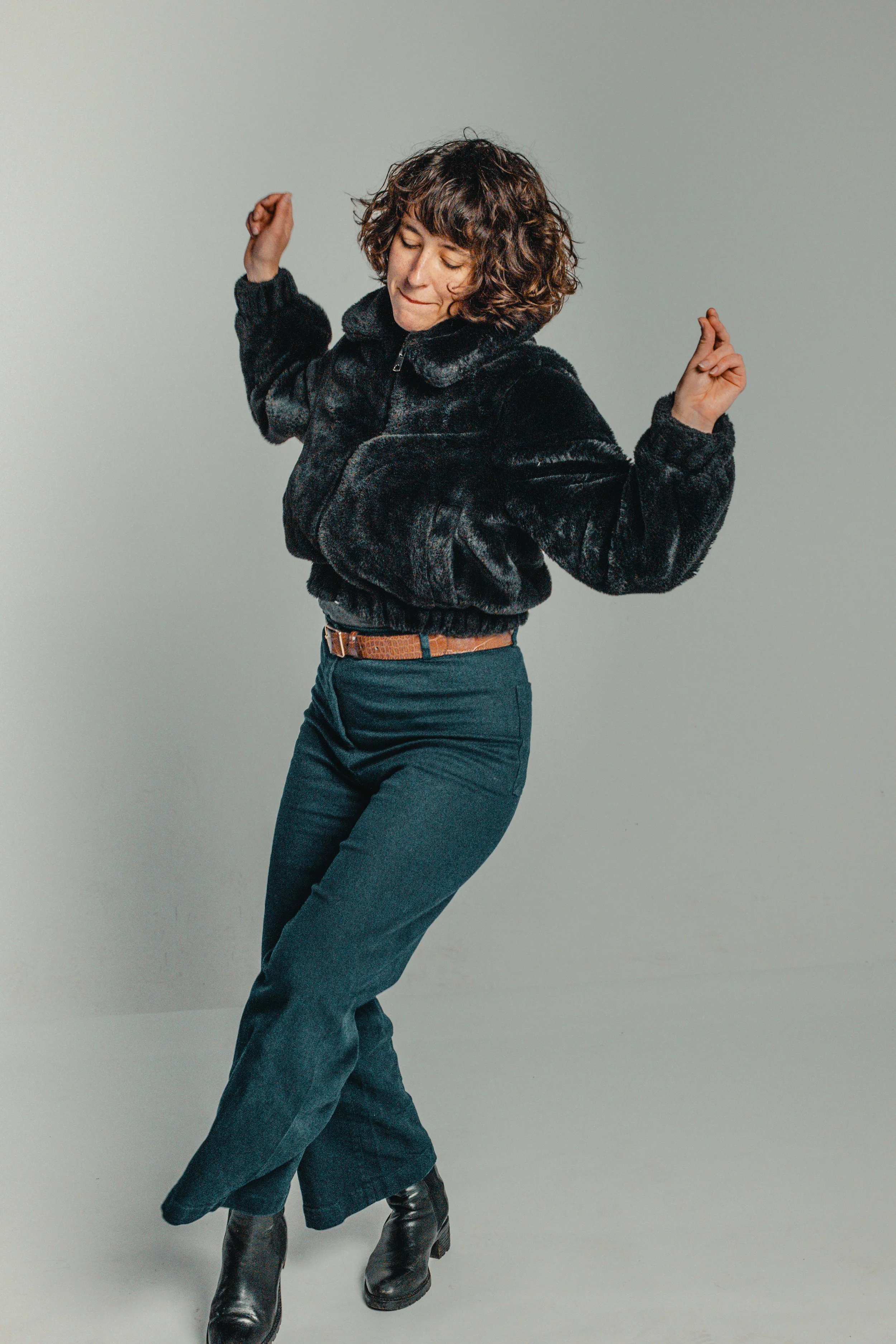 Woman dancing in a black fuzzy jacket, dark jeans, and black boots against a neutral background.