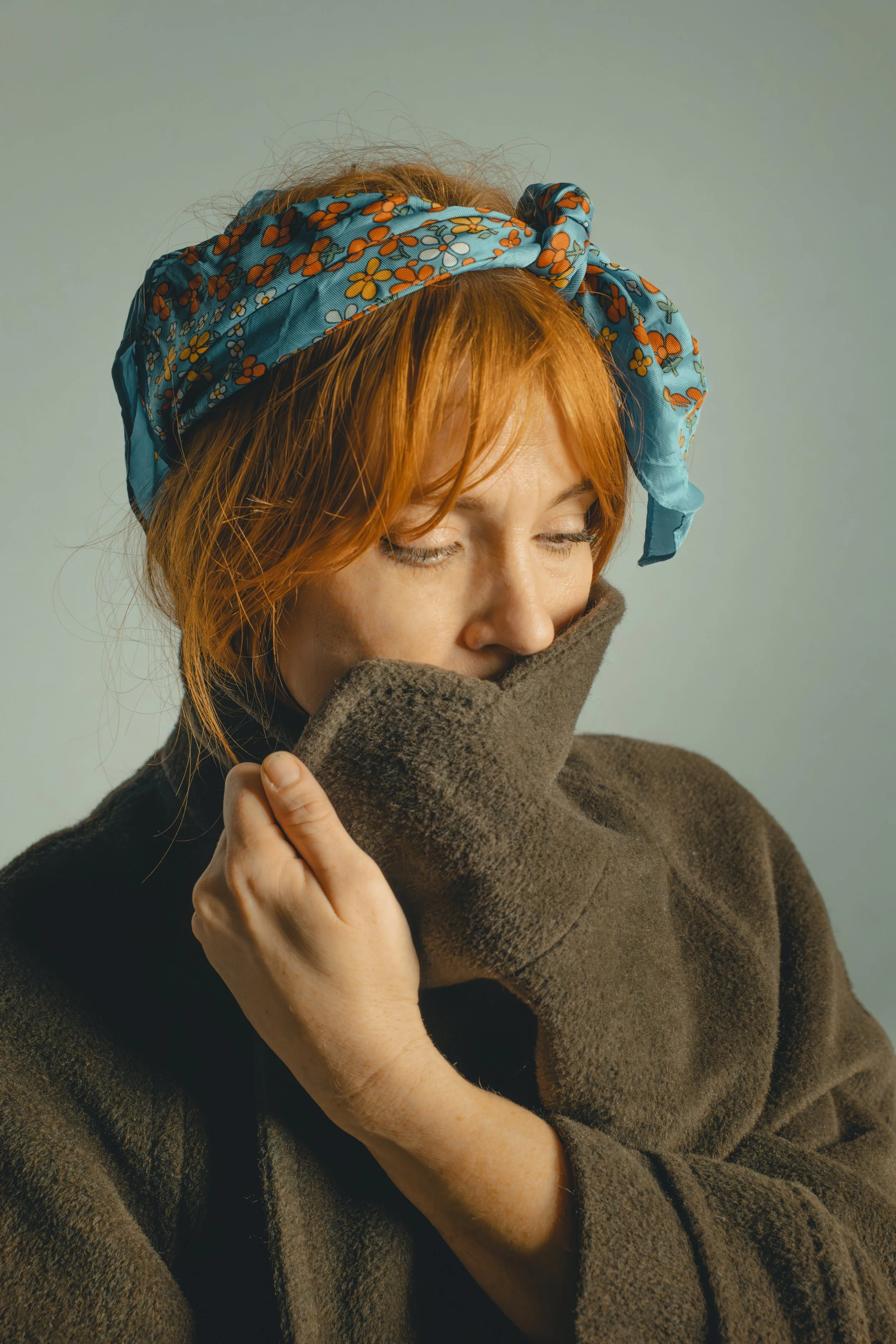 A woman with red hair wearing a blue floral headscarf and a brown coat, holding the collar of her coat up to her face with her eyes closed.