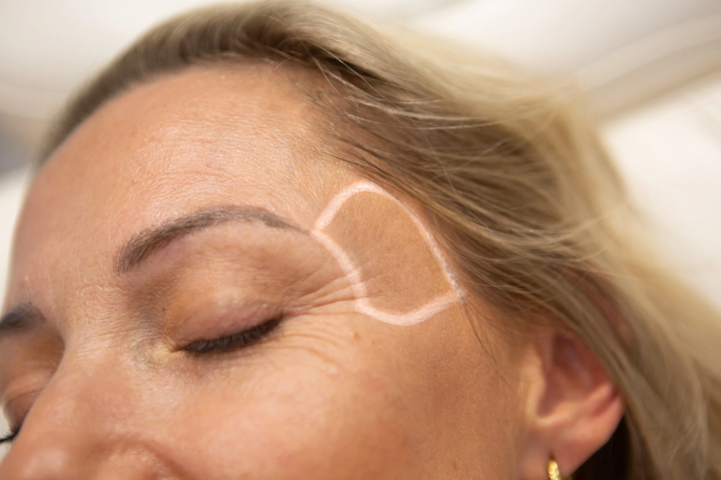 Close-up of a woman with makeup on her face and a shape drawn on her temple for cosmetic tattooing.