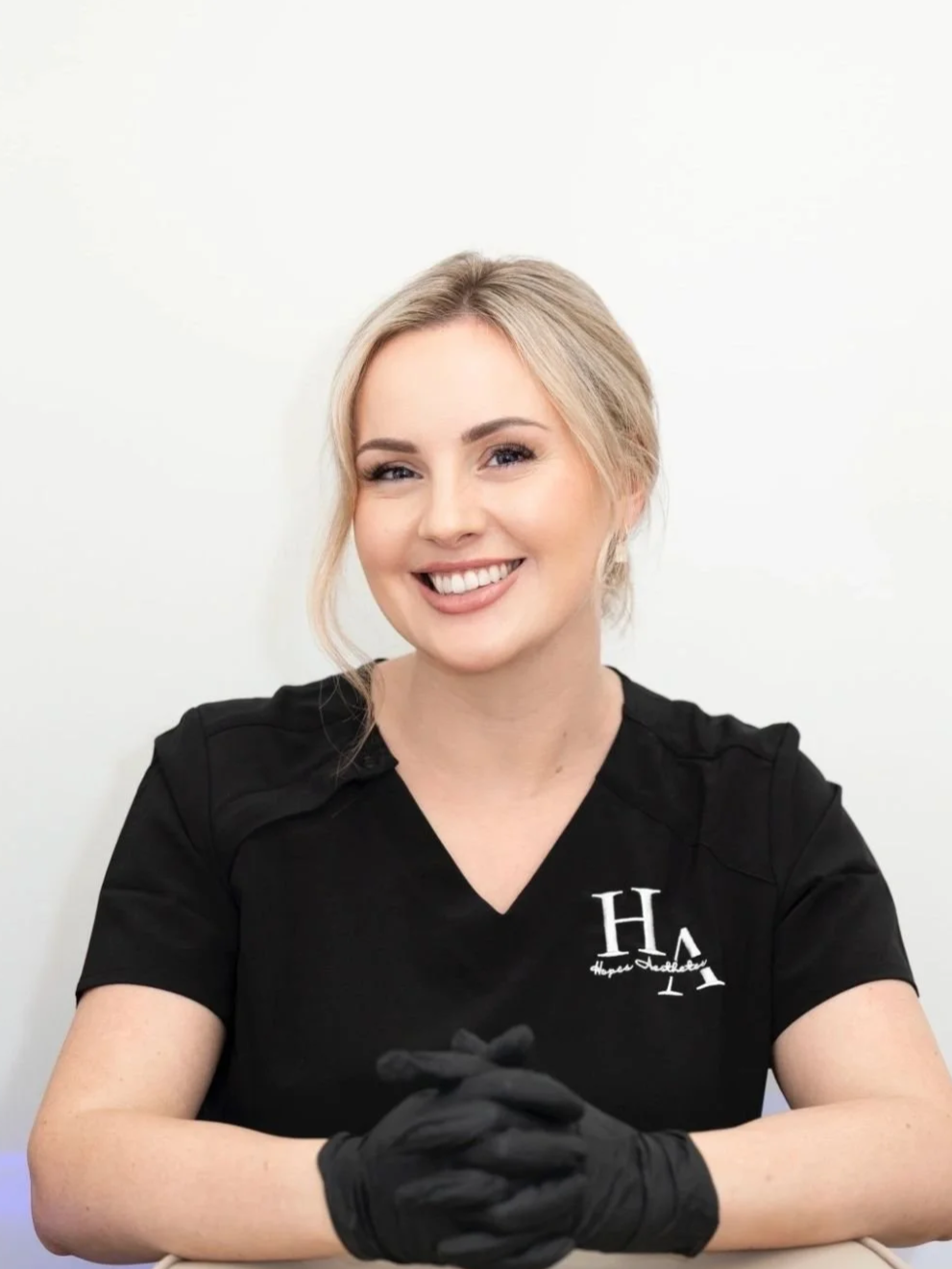 A smiling woman with blonde hair wearing a black shirt with white writing, sitting with hands clasped in front of her, against a plain white background.