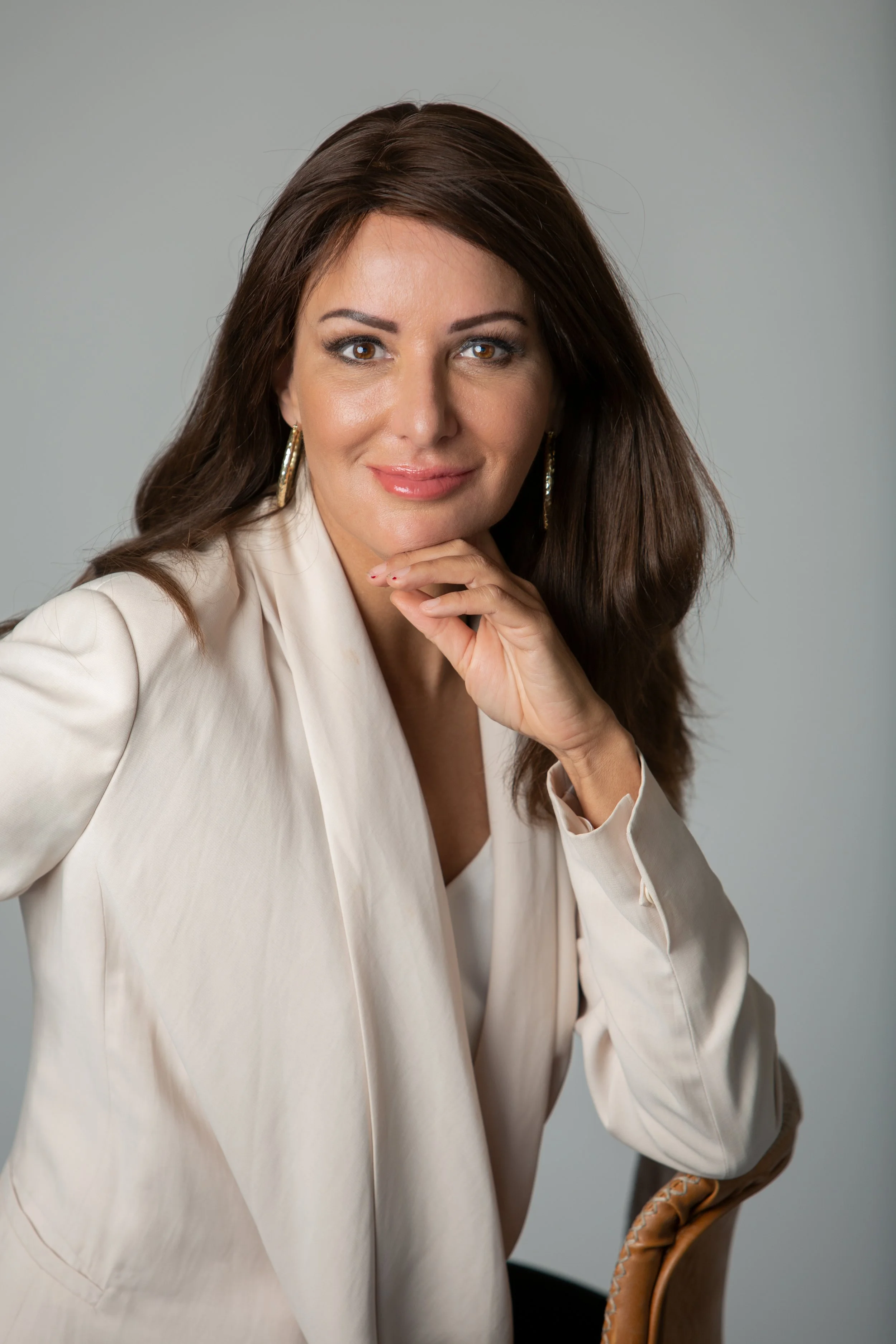 A woman with long brown hair, wearing a cream blazer, seated with her chin resting on her hand, looking at the camera with a slight smile, against a plain gray background.