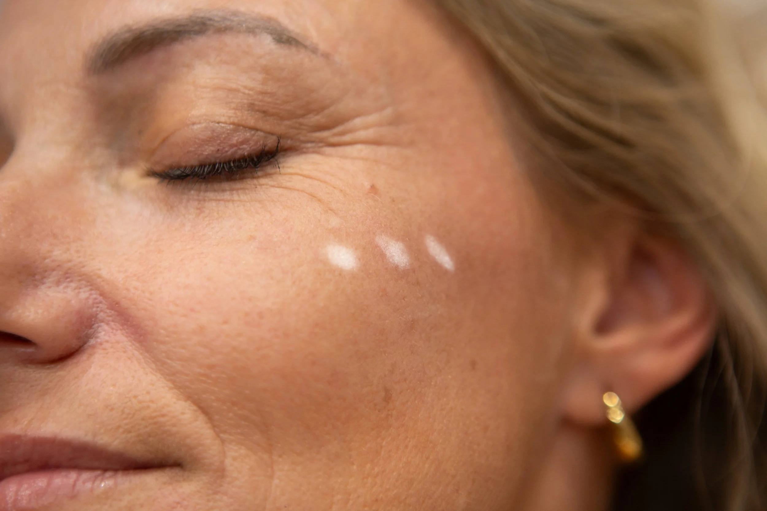 Close-up of a woman's face with her eyes closed, showing makeup, skin texture, and a small earring.