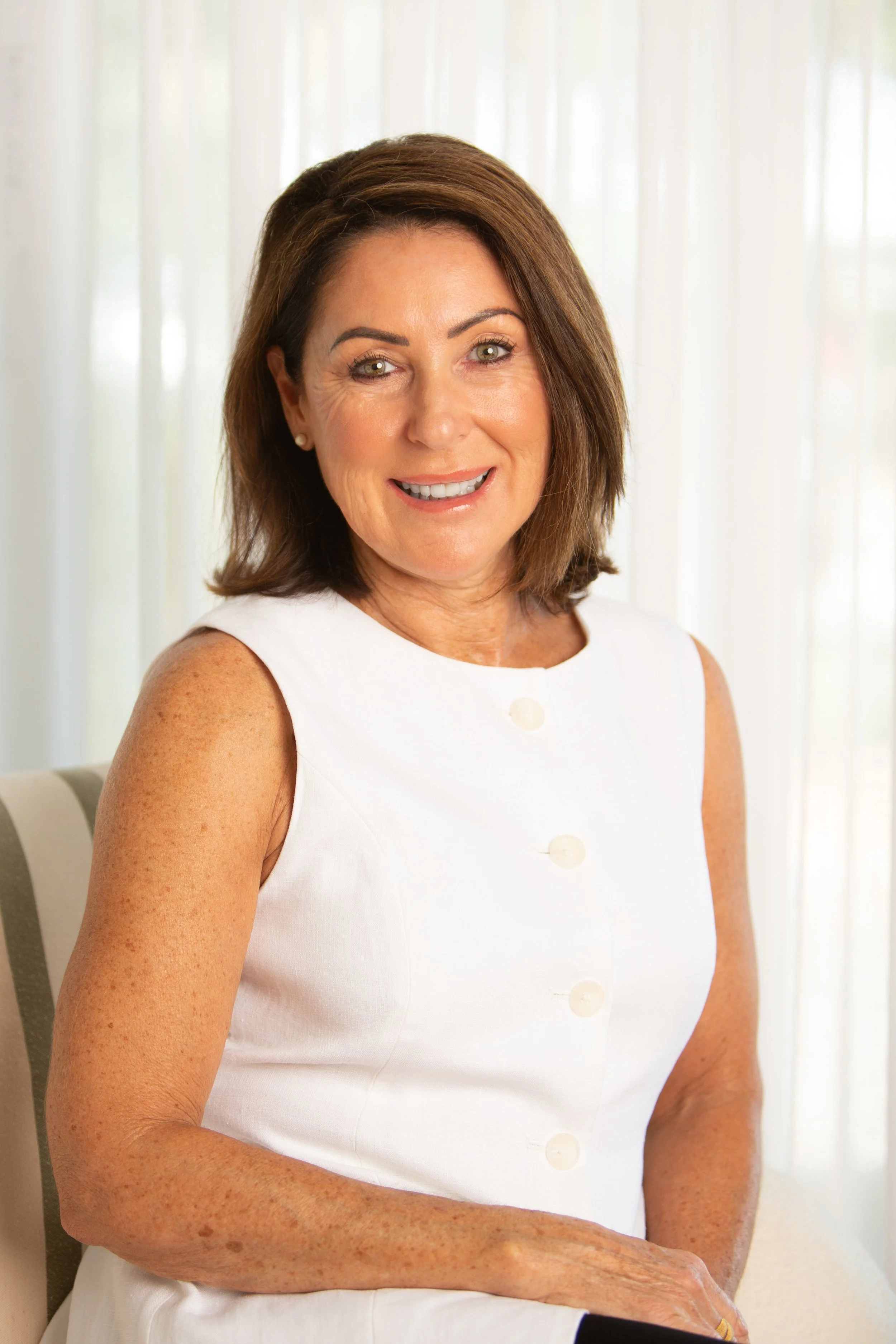 A woman with shoulder-length brown hair and light skin, smiling while sitting indoors in front of white curtains, wearing a sleeveless white dress with buttons.