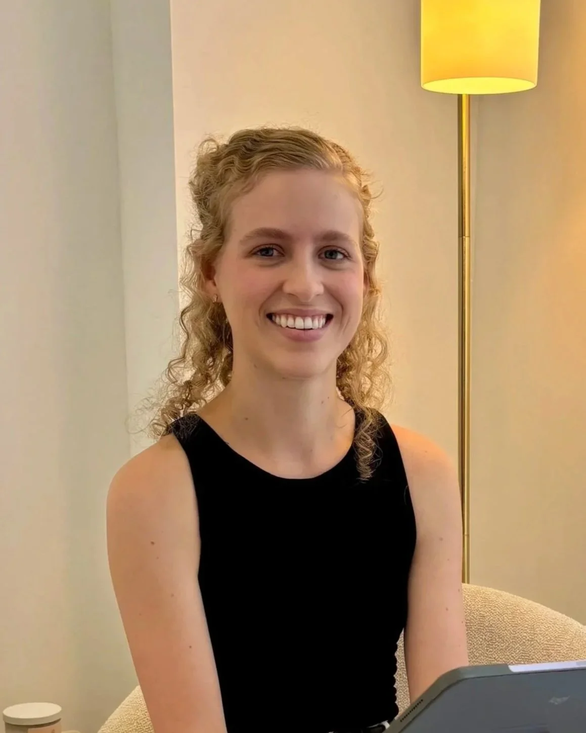 A woman with curly blonde hair smiling, wearing a black sleeveless top, seated in a room with a lamp behind her.