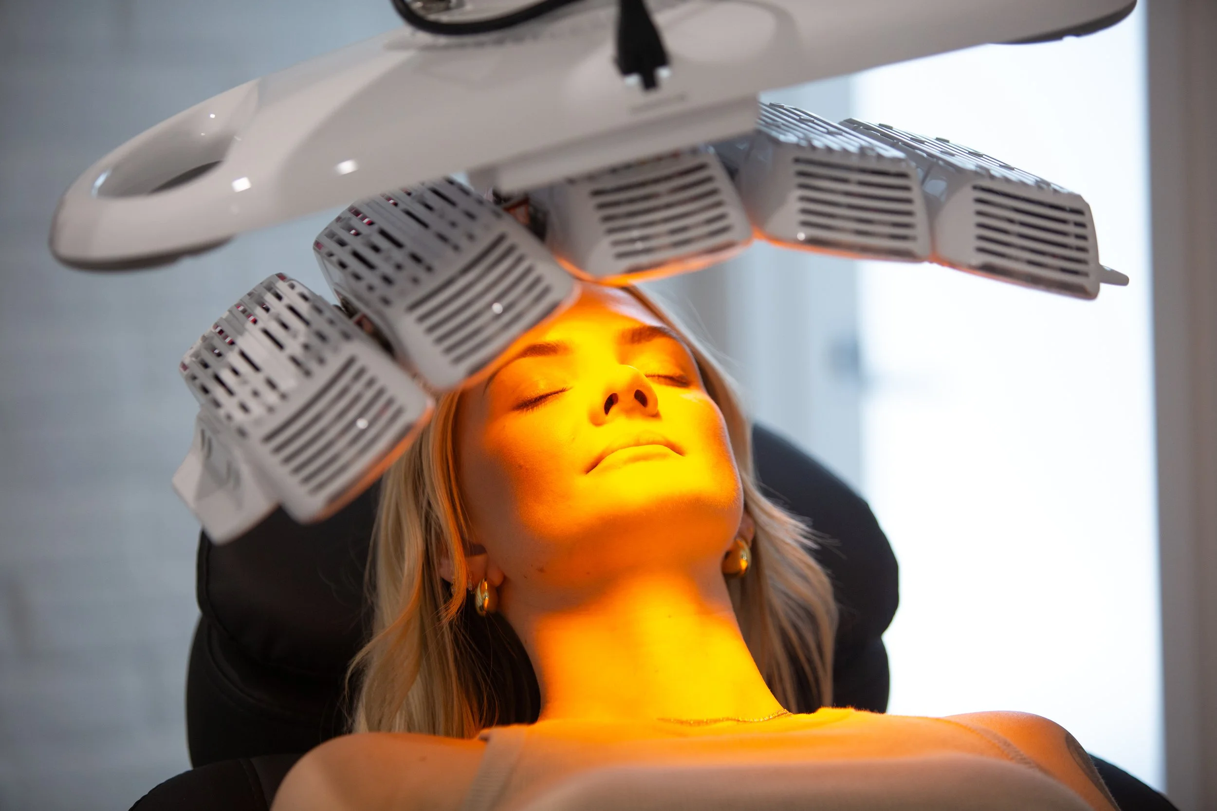 A woman receives a facial treatment under a red light therapy device with multiple lamps in a spa or clinic setting.