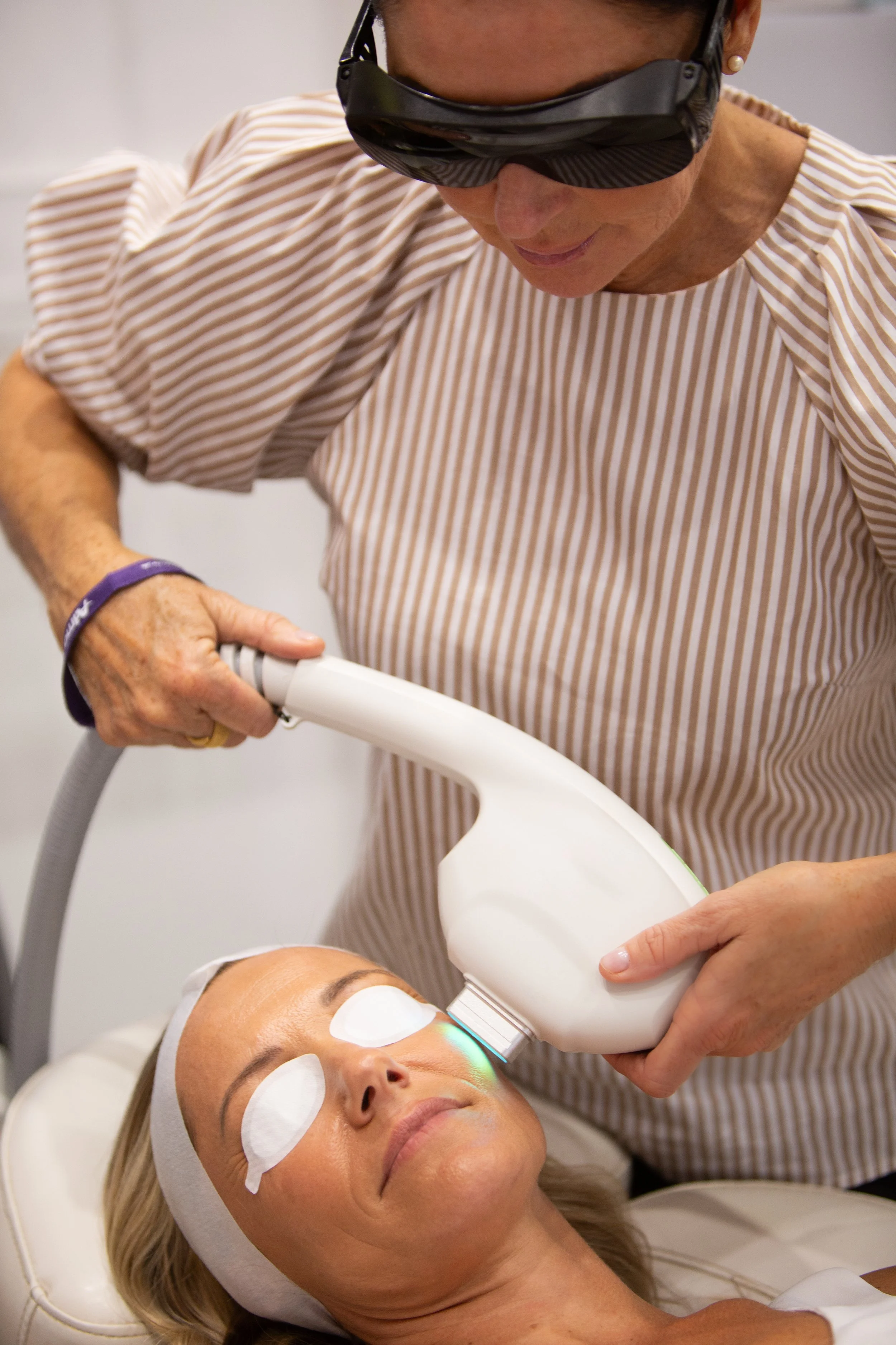 A woman receiving a facial treatment with a handheld device at a spa or clinic, with eye pads and a headband, while a technician uses a specialized machine.