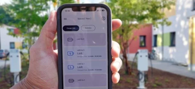 Nahaufnahme einer Hand, die ein Smartphone mit einer App zur Überwachung von elektrischen Ladestationen hält, im Hintergrund Bäume und bunte Gebäude.