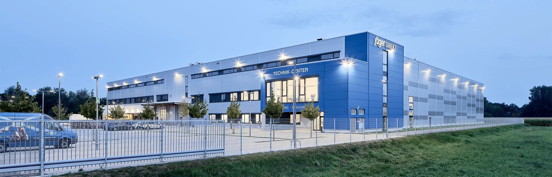 Moderne blaue Industriehalle mit Parkplatz und Umzäunung bei Dämmerung