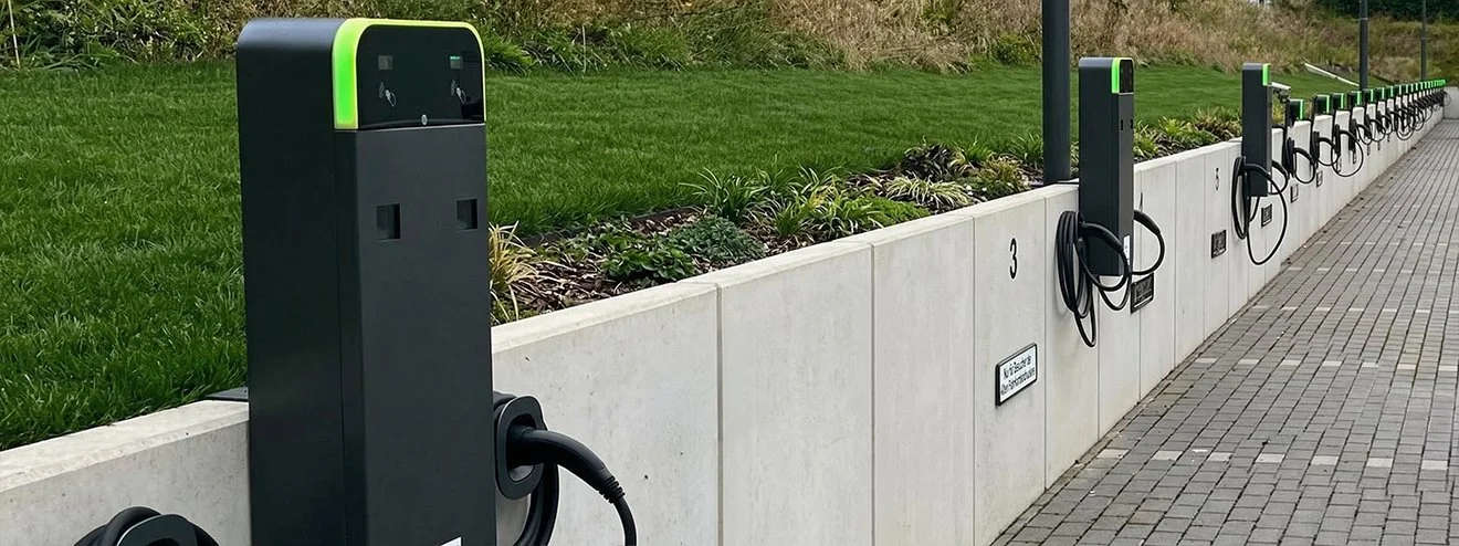 Elektroladestationen für E-Bikes in einer Reihe an einem Gehweg, daneben eine bepflanzte Grenze mit Gras und Pflanzen.