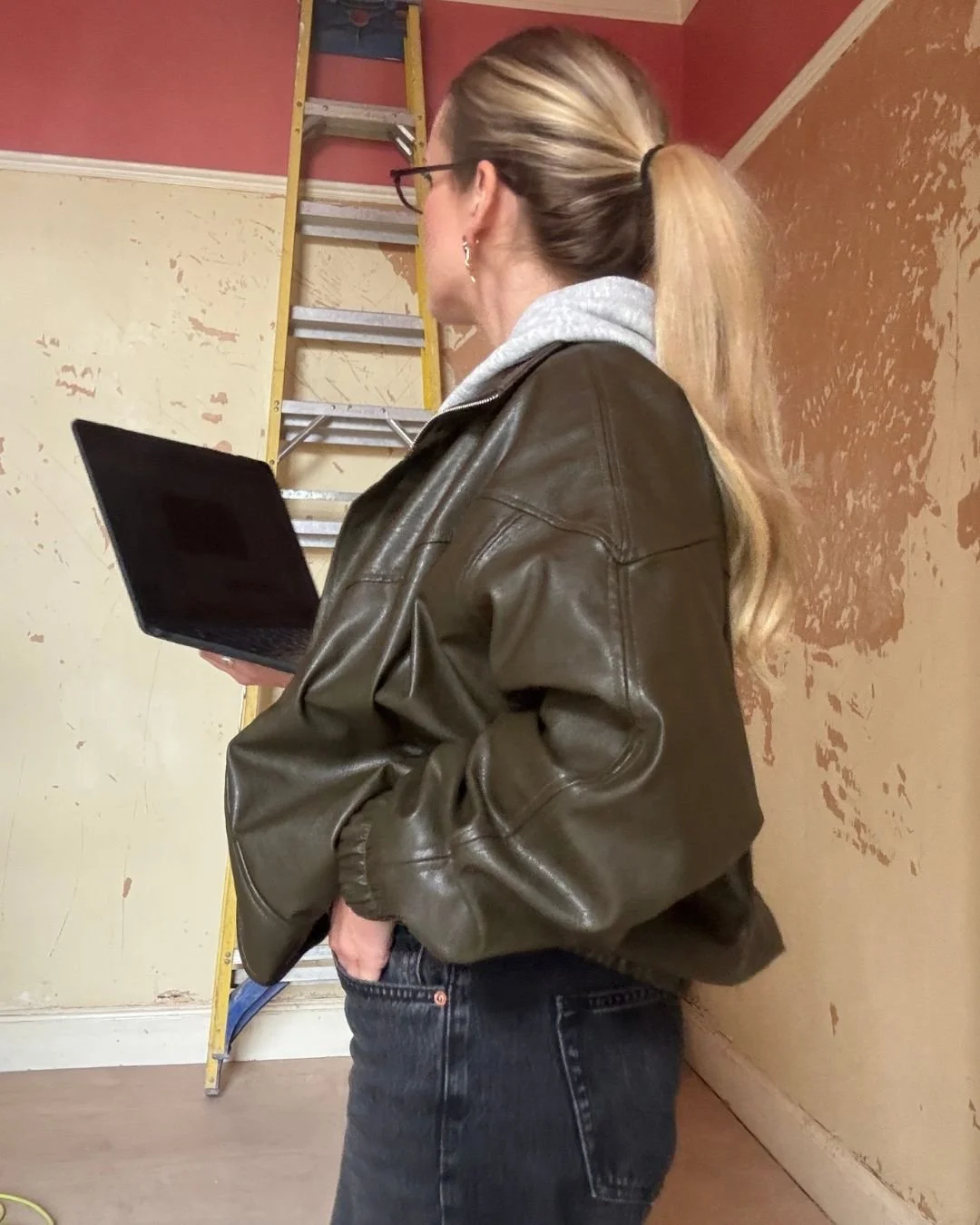 A woman with blonde hair tied back, wearing glasses, a gray hoodie under a brown leather jacket, and black jeans, holding a laptop in her right hand, standing in front of a partially painted wall with a yellow ladder in the background.