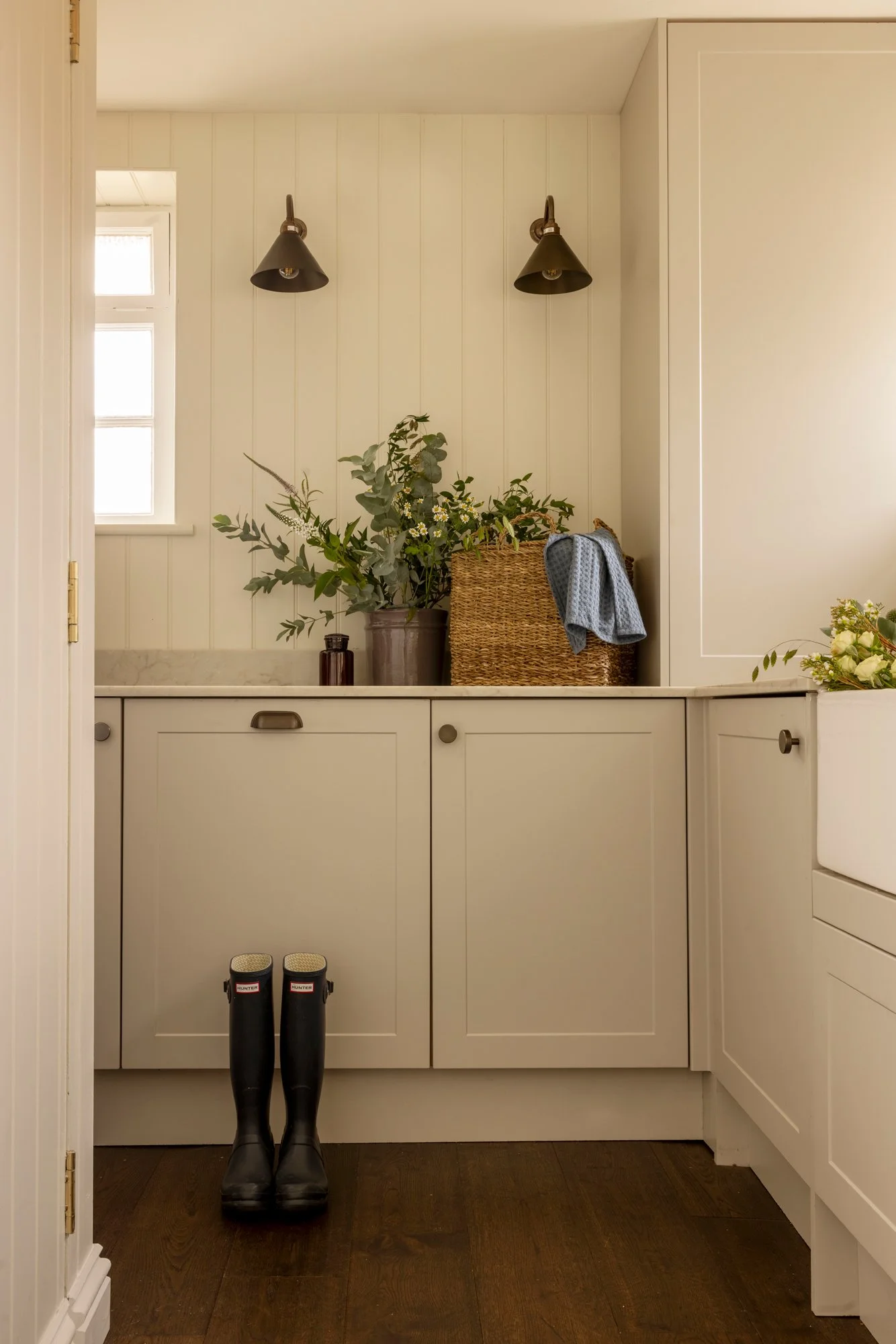 A view of a small entryway or mudroom with white paneled cabinetry and dark wooden flooring. There are two tall windows on the upper left bringing in natural light. On the countertop, there is a large potted plant, a small dark bottle, and a woven ba