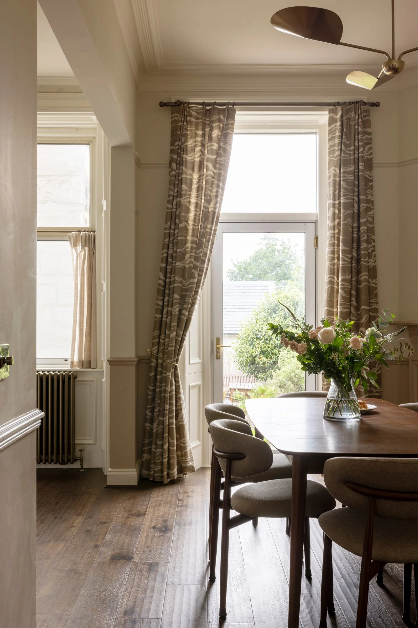 Bright dining room with large window, beige curtains, wooden table with a glass vase filled with flowers, and upholstered chairs.