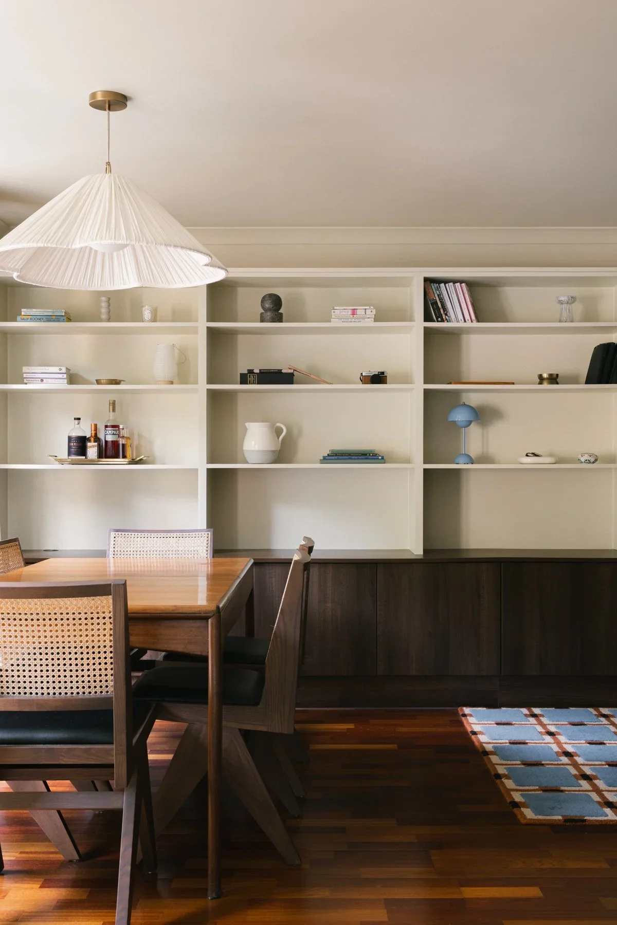 Living room with white built-in bookshelf, wooden dining table, black chairs, a white pendant light, and a multicolored rug.
