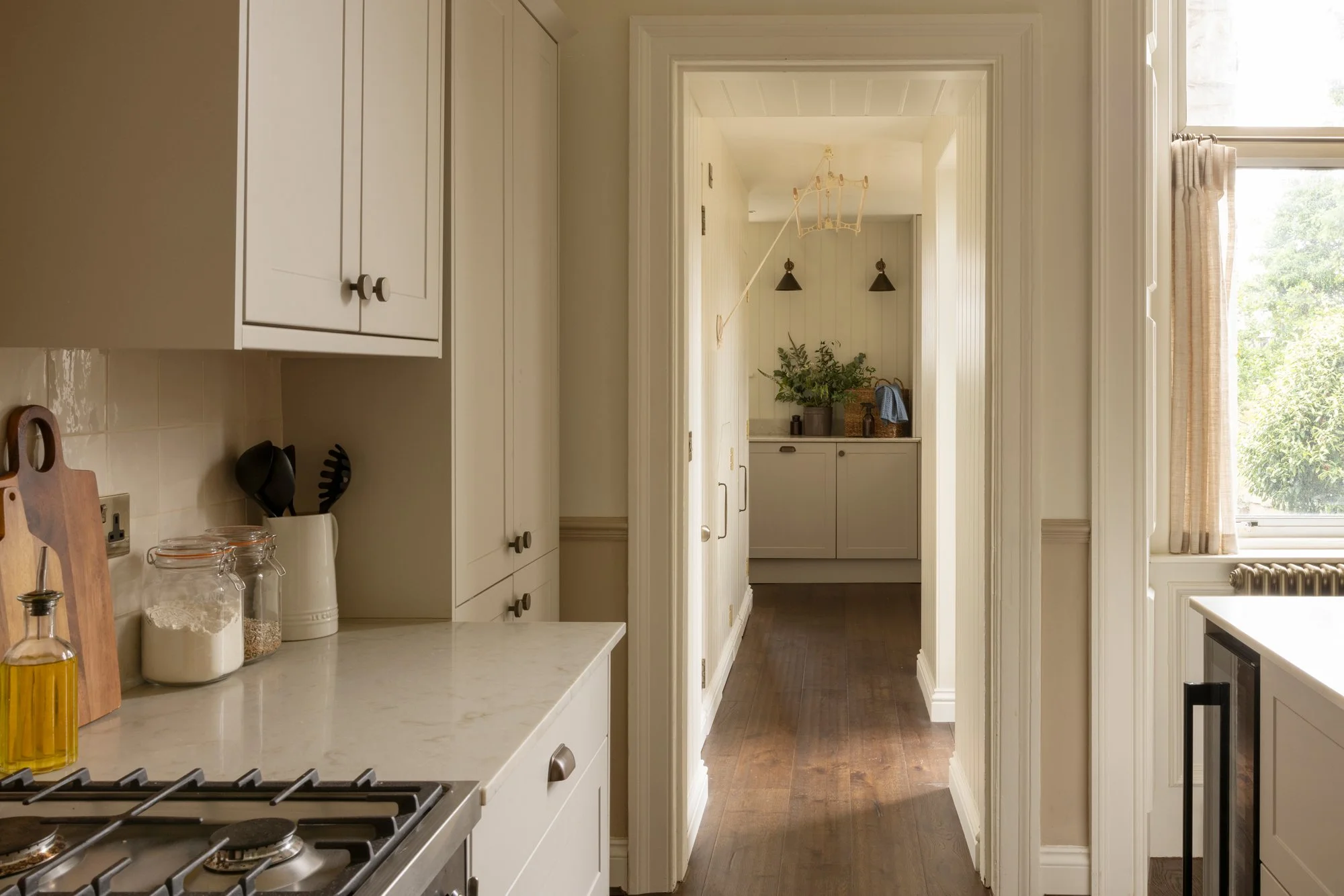 A view from a kitchen looking into a hallway with a cozy and bright atmosphere. The kitchen features white cabinets, a marble countertop, and glass jars containing baking ingredients. There's a gas stove and a window with beige curtains letting in na