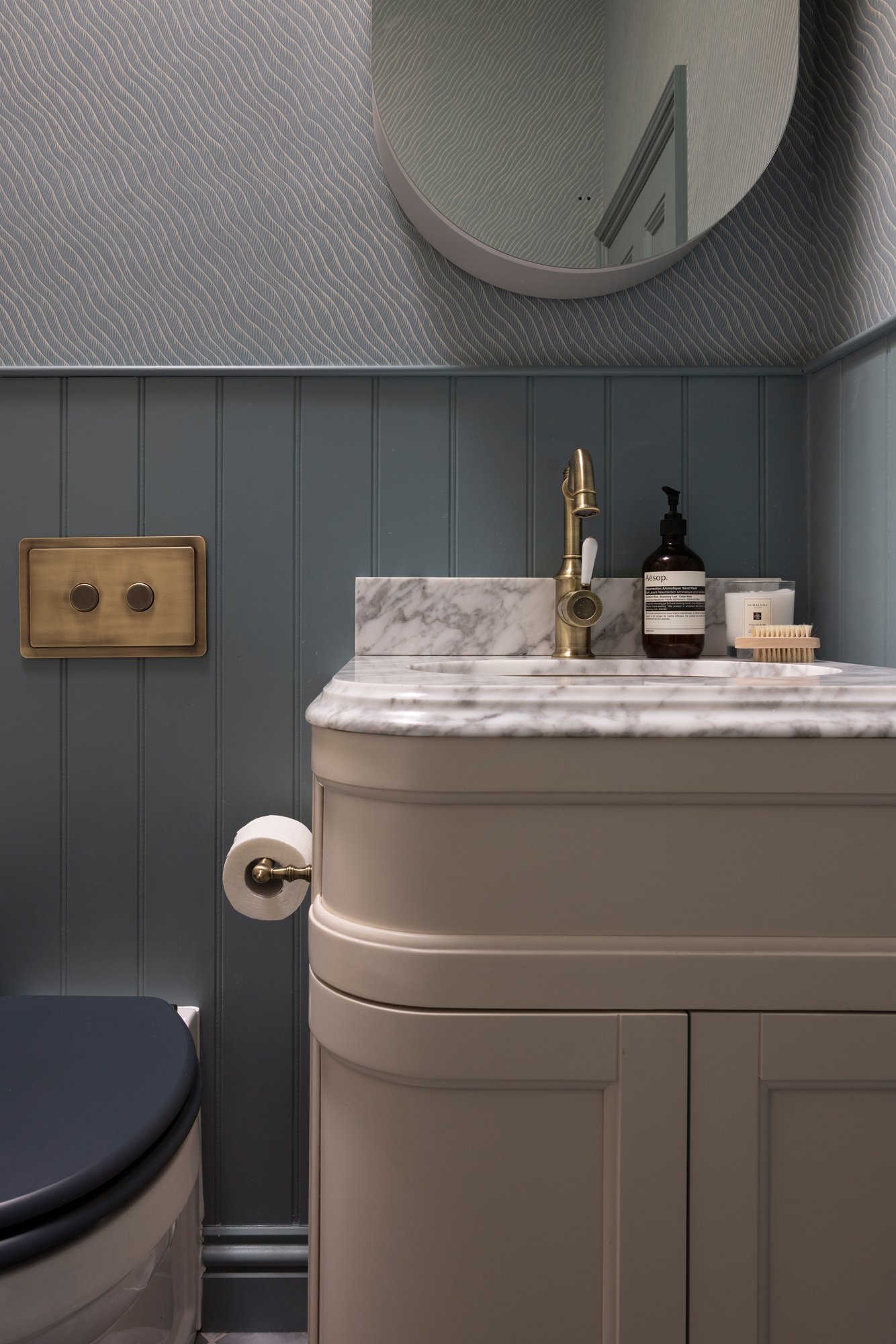 Small bathroom with a marble countertop sink, gold faucet, mirror, Aesop hand soap, a container with a brush, toilet paper on a holder, and a partially visible toilet and blue wall paneling.