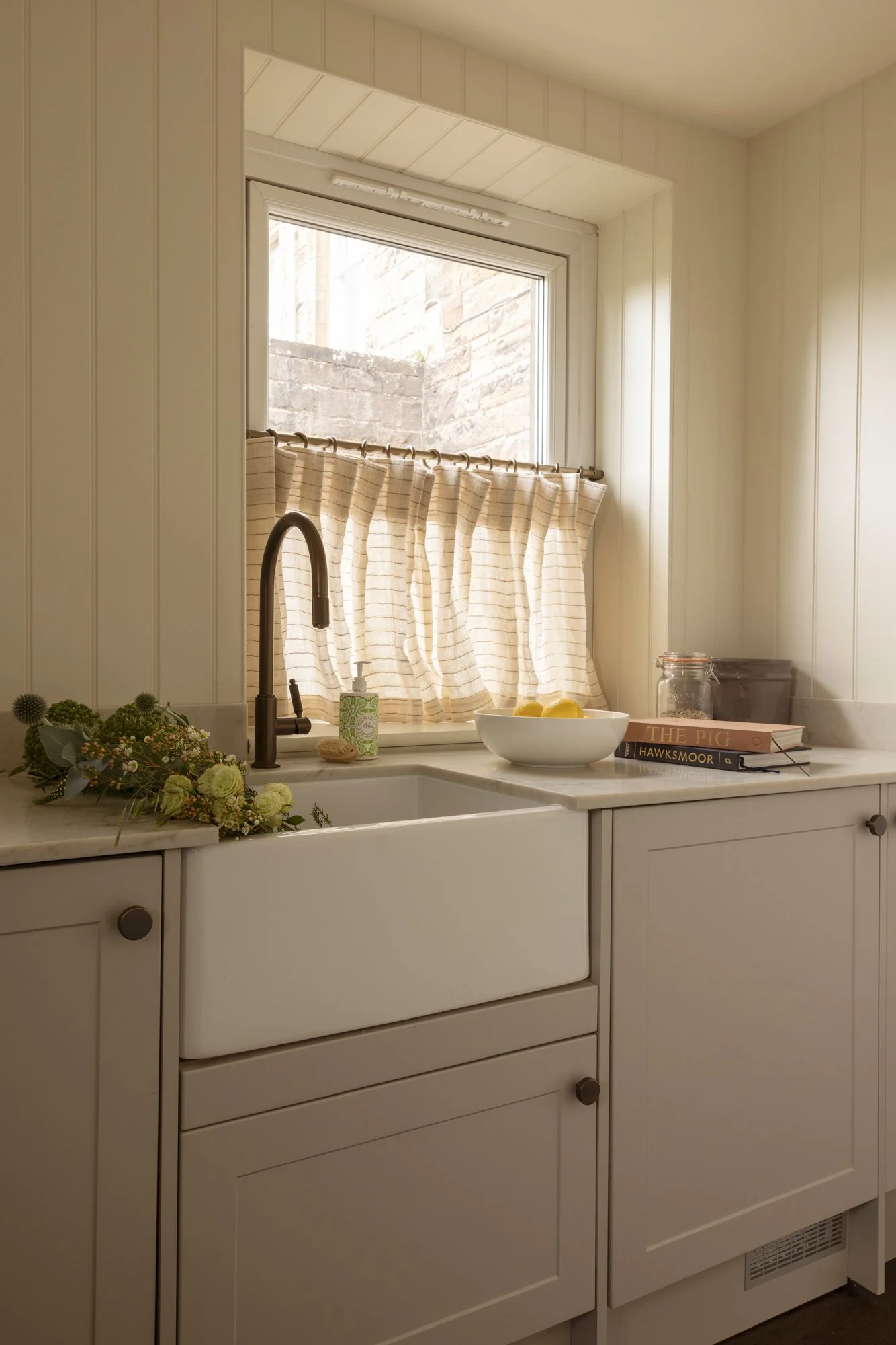 A kitchen window with beige striped curtains above a white farmhouse sink. On the countertop, there are books, a glass jar, a bowl of lemons, and a floral arrangement.