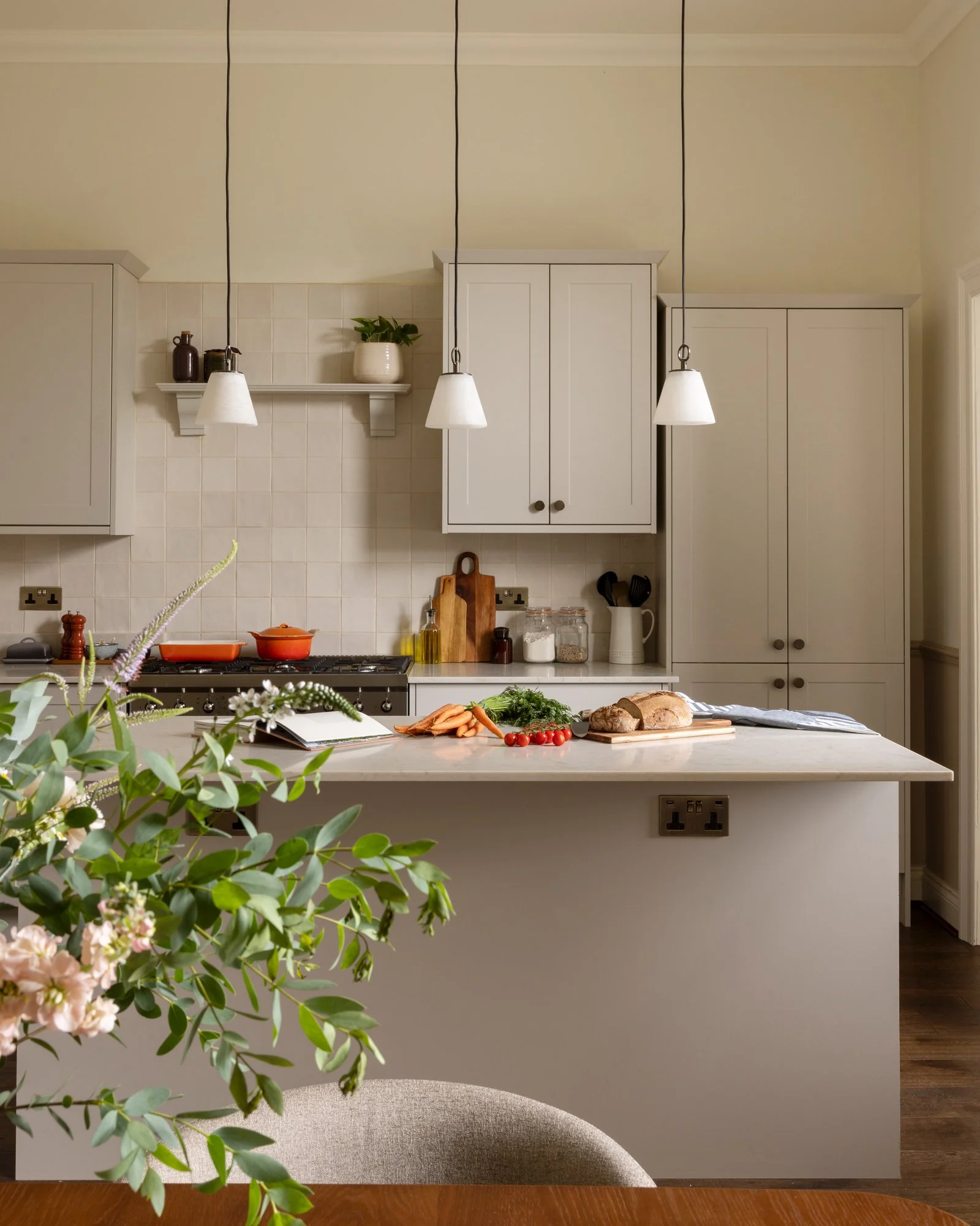 A kitchen with white cabinets, a white tiled backsplash, and a marble kitchen island. On the island are fresh carrots, cherry tomatoes, leafy greens, a loaf of bread, and a book. Hanging above are three white pendant lights. There are jars and cuttin