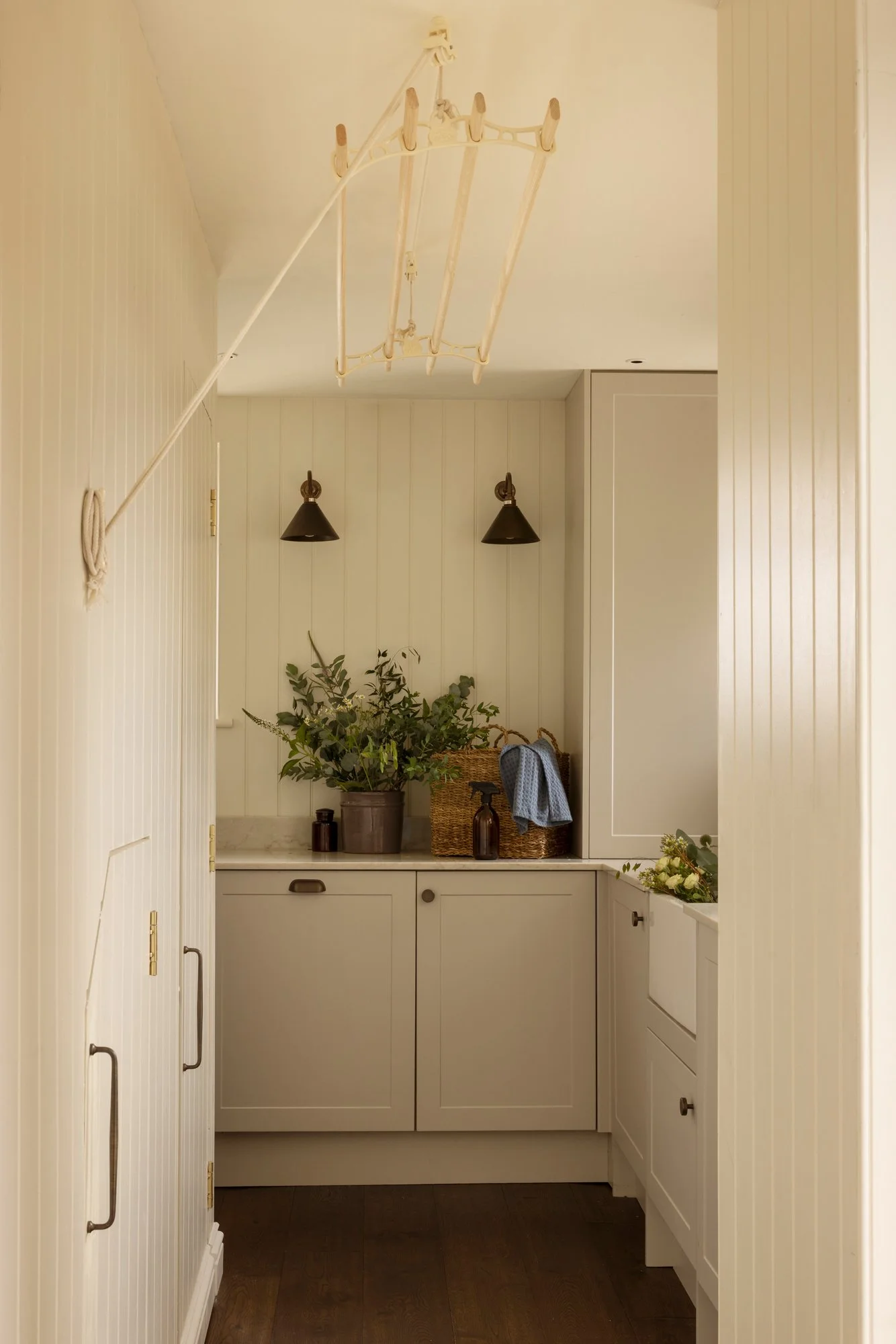 A cozy kitchen nook with white paneled walls and cabinetry. Decorative items include potted plants, a woven basket, and a few small bottles. A wall-mounted light fixture with two shades illuminates the area, with a blue cloth hanging on the cabinet h