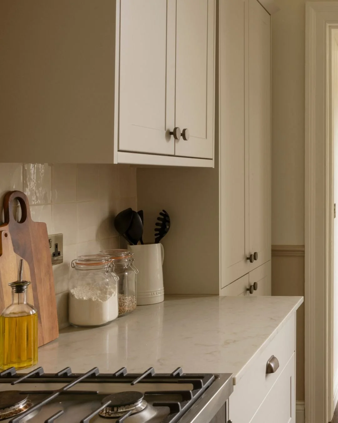 A kitchen countertop with jars of flour, rice, and oats, a wooden cutting board, a bottle of olive oil, a set of black cooking utensils in a white holder, and cabinets overhead.