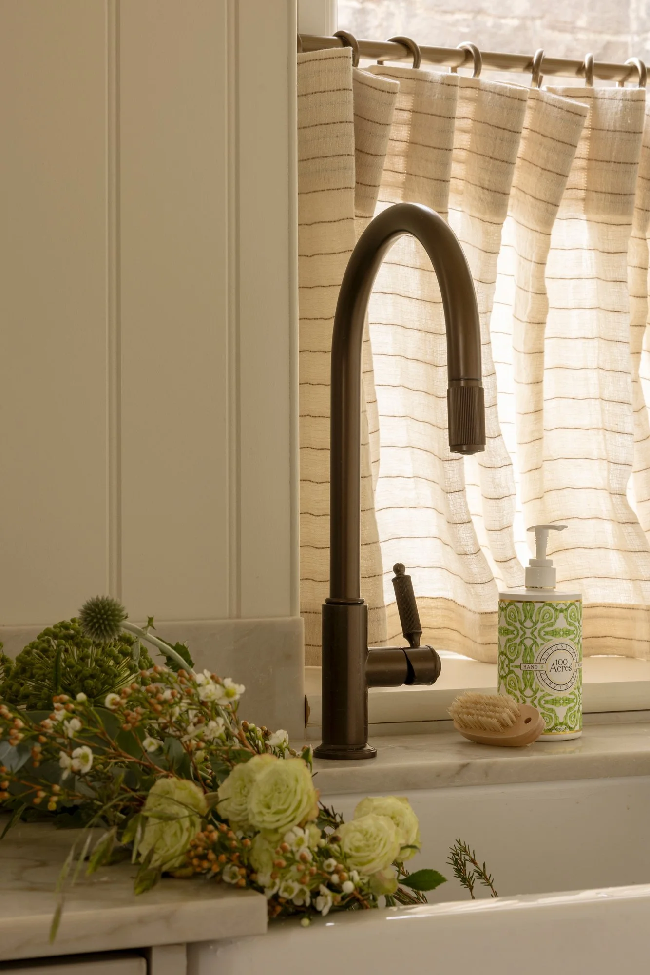 A kitchen sink with beige striped curtains in the background. The sink has a dark bronze faucet, a bar of soap, a green and white decorative hand soap dispenser, and some floral arrangements on the countertop.