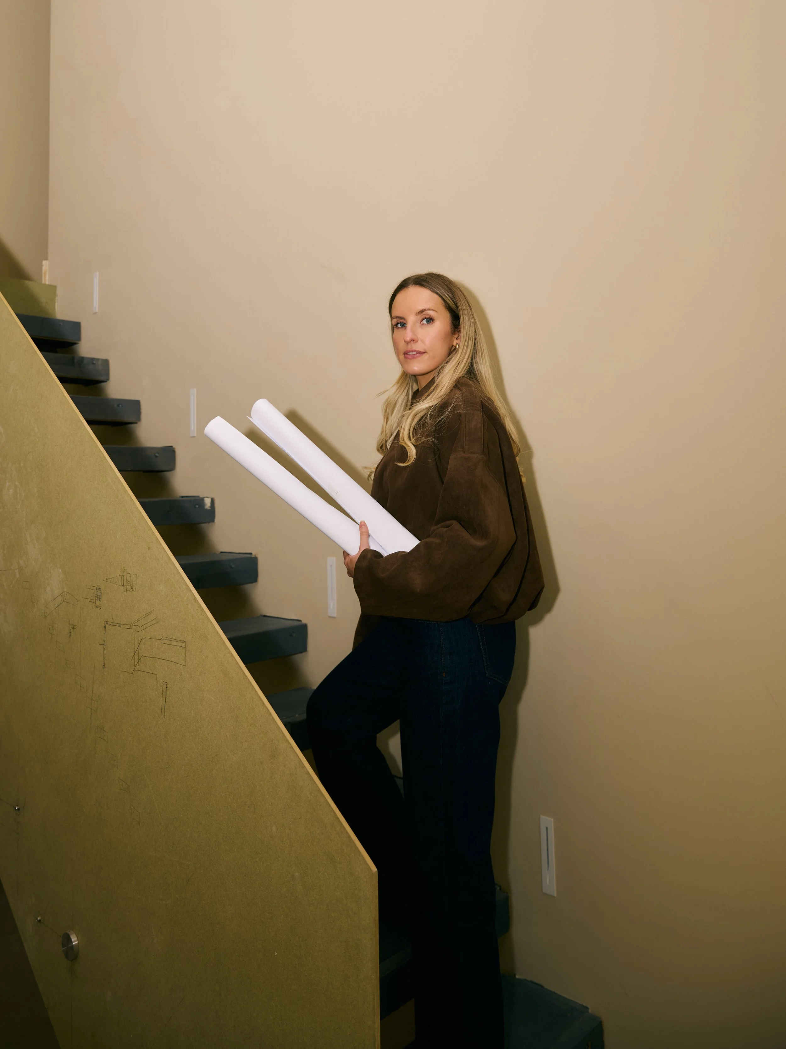 Courtney holding rolled-up blueprints standing on staircase inside a building.