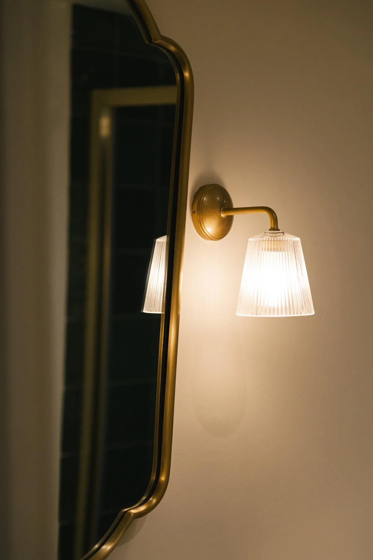 Close-up of a wall-mounted brass sconce with a pleated glass lampshade, illuminating a beige wall with a subtle shadow.