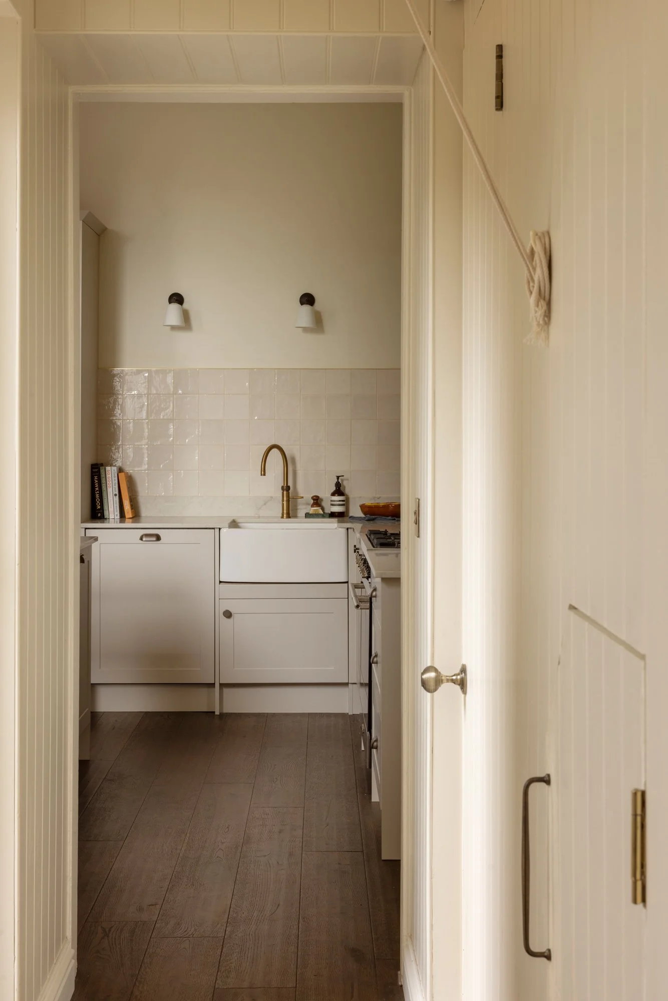 View of a small kitchen seen through a doorway with cream-colored walls, white cabinets, a gold faucet, and a white tile backsplash. There are two books on the counter on the left and kitchen essentials on the right, with dark wood flooring.