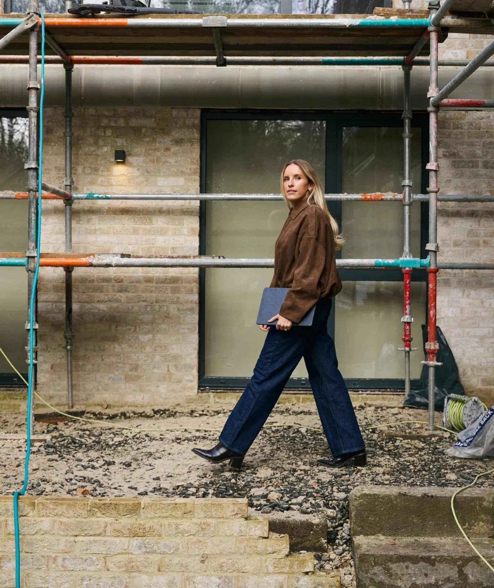 A woman dressed in a brown jacket and dark blue pants is walking past a construction site with metal scaffolding and a brick building in the background, holding a black folder or laptop.