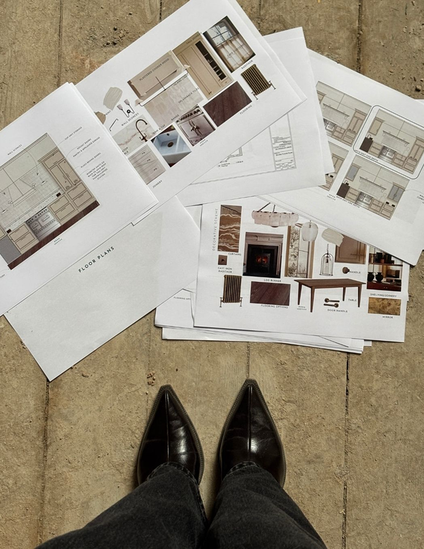 A person's feet wearing black leather shoes and dark pants, standing on a concrete floor, looking down at scattered architectural and interior design plans and mood boards.