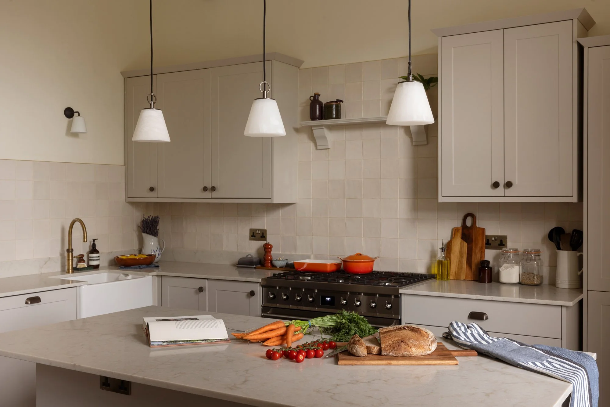 A modern kitchen with white cabinets and a marble countertop, featuring a gas stove with red cookware, fresh vegetables, and a book open on the counter.