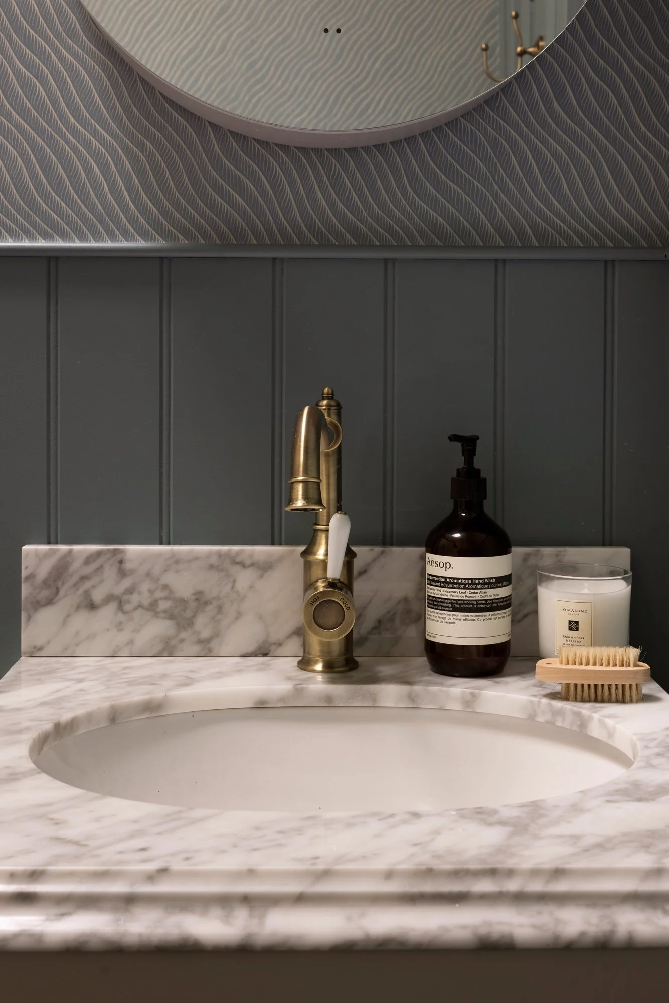 Bathroom sink with a marble countertop, a gold faucet, a brown Aesop hand soap bottle, a white candle, and a small wooden hairbrush, with dark gray paneled wall and a round mirror above.