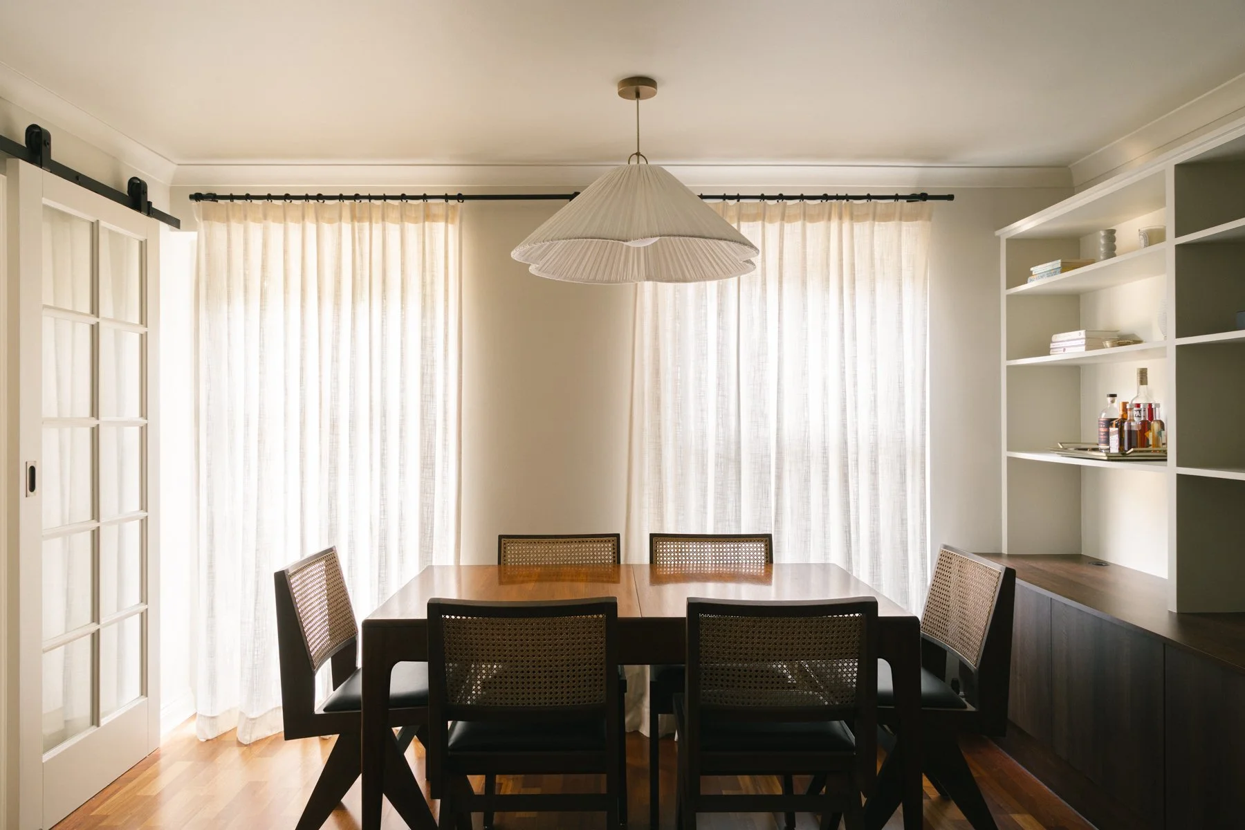 Dining room with a wooden table, six chairs with cane seats, cream curtains, a white ceiling, a hanging lampshade, a sideways sliding door, and a white built-in bookshelf with books and bottles.