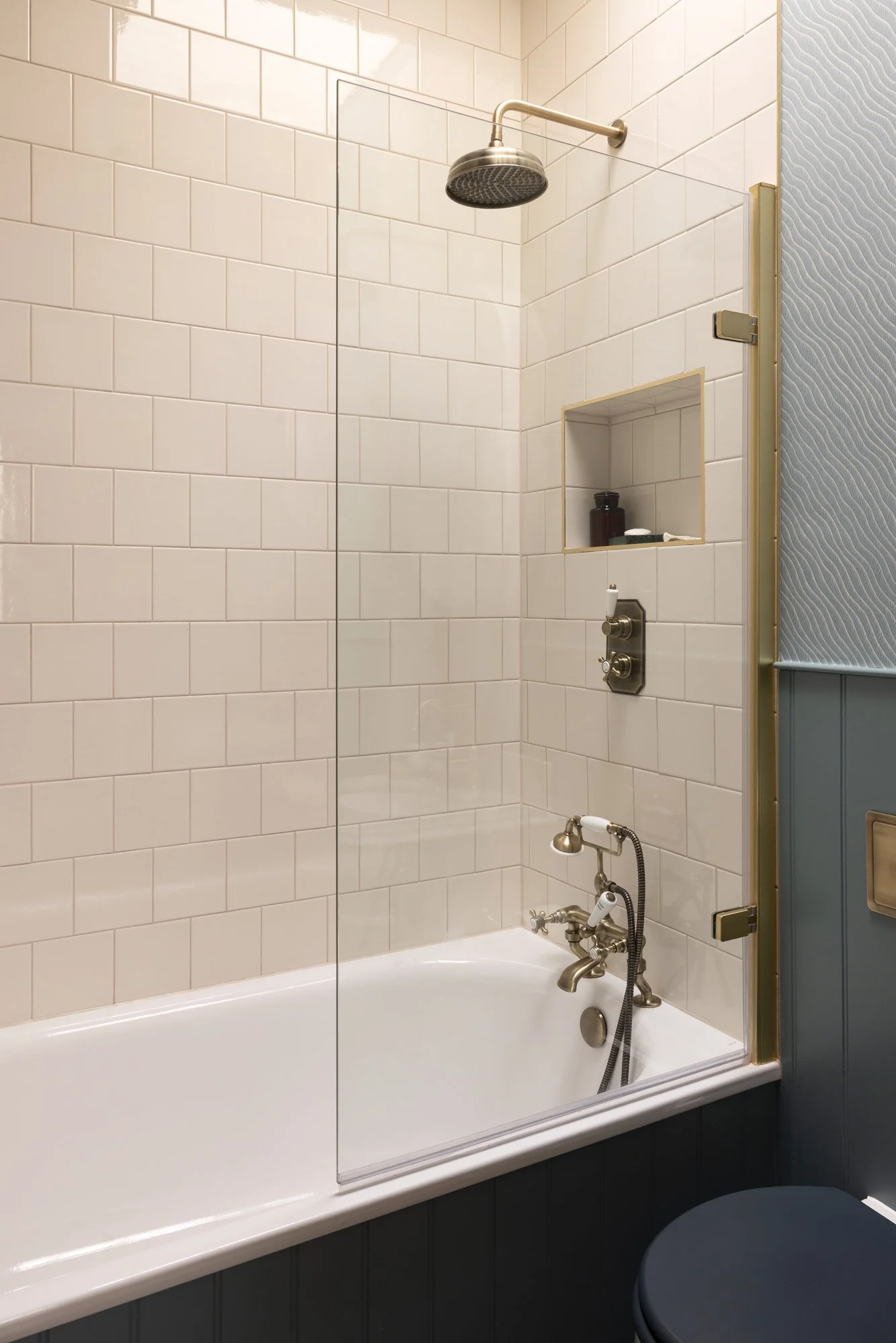 Bathroom shower with white tiled walls, a stainless steel showerhead, a built-in shelf with bottles, and a bathtub at the bottom. The shower area is partially enclosed with a glass partition, with a textured blue wall on the side.